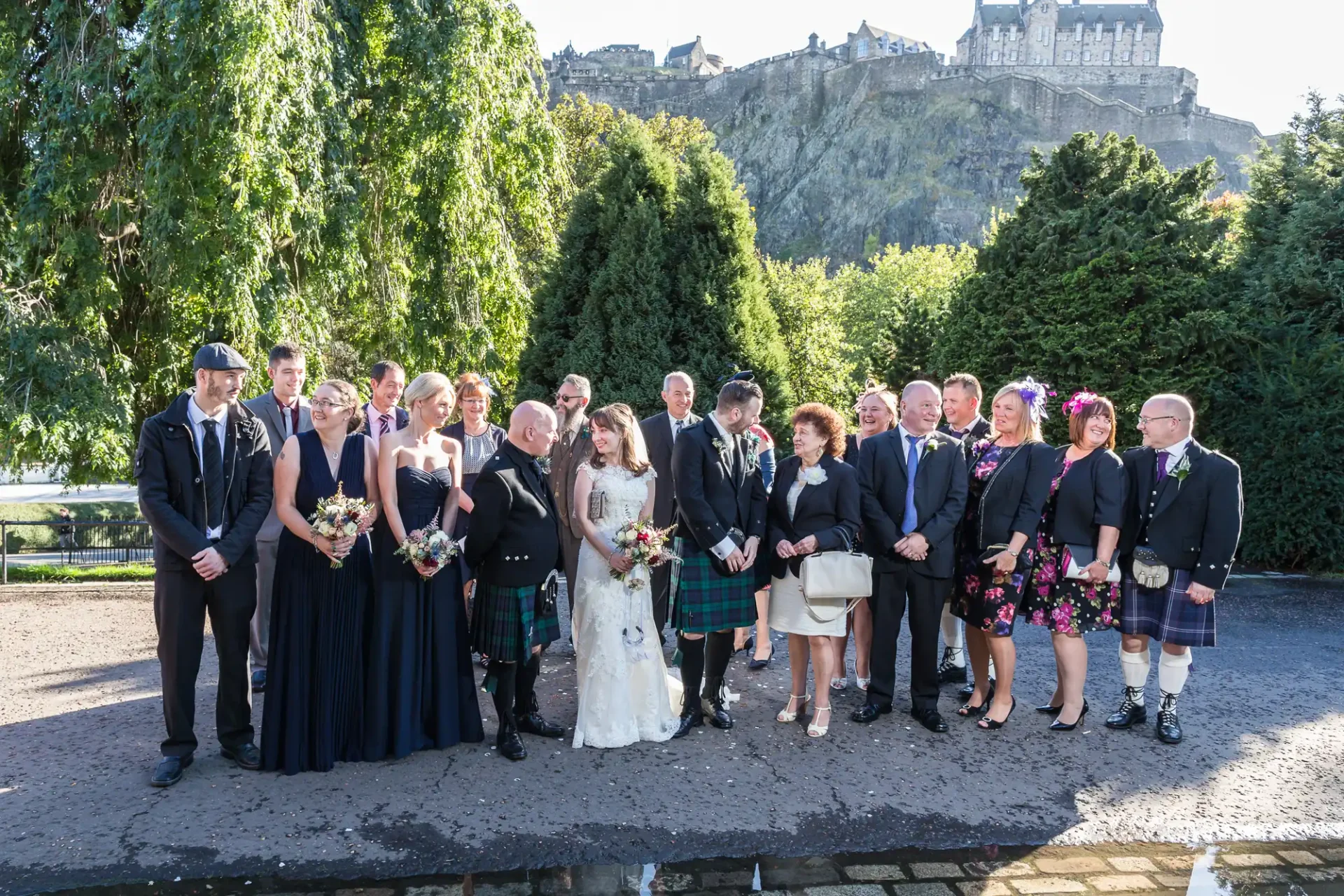 Wedding group photo with the bride, groom, and guests in formal attire, some in kilts, with edinburgh castle in the backdrop.