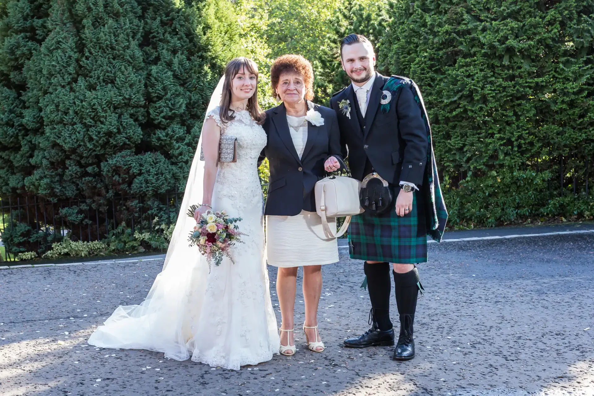 A bride in a white gown, a groom in a kilt, and an older woman in a cream dress posing together outdoors, with a backdrop of dense green shrubs.