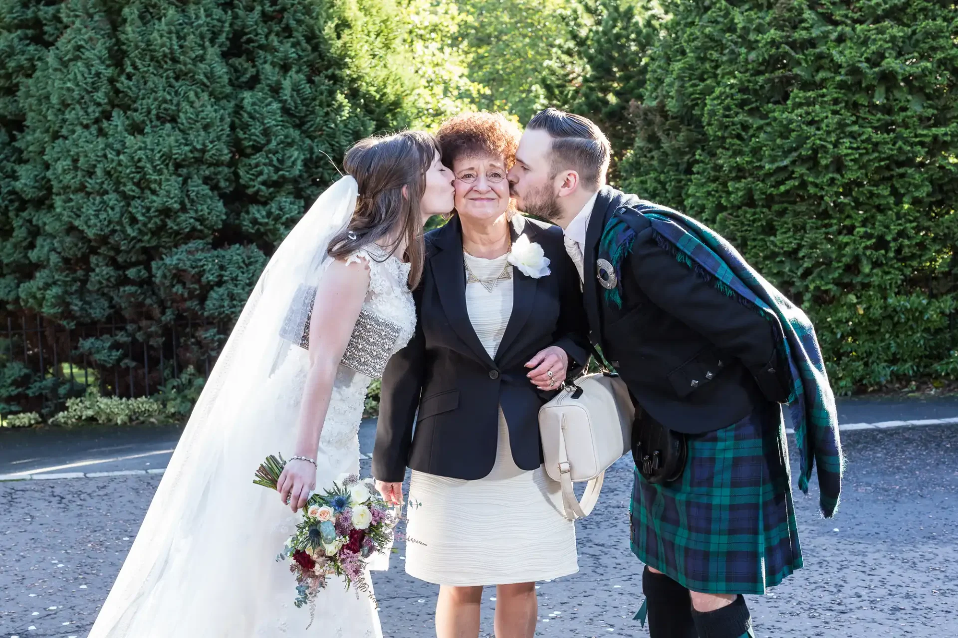 A bride and groom in wedding attire affectionately kiss an older woman on her cheeks outside, with trees in the background. the groom wears a kilt.