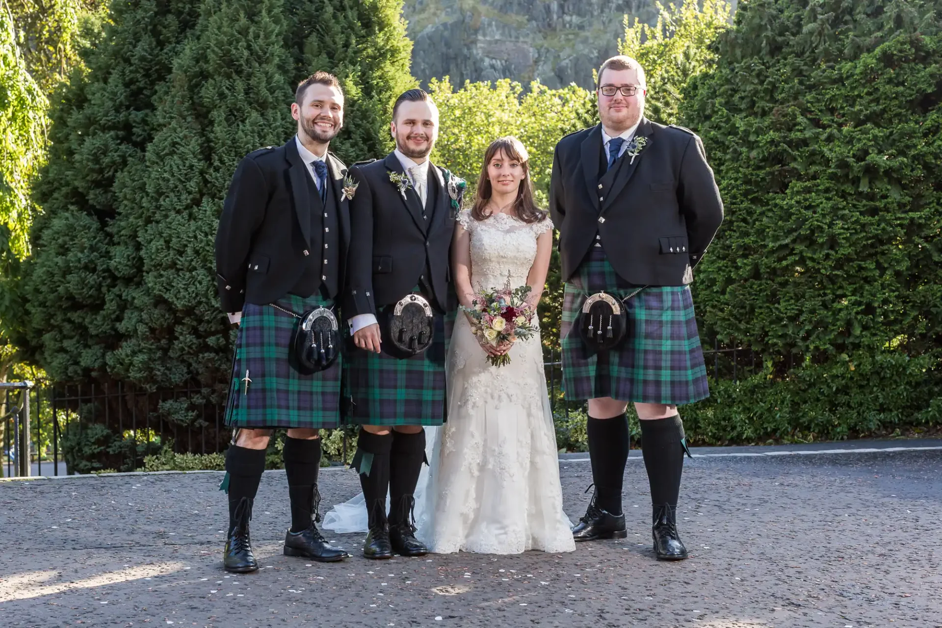 A bride in a white lace gown and three men in traditional scottish kilts and jackets, posing together outdoors.