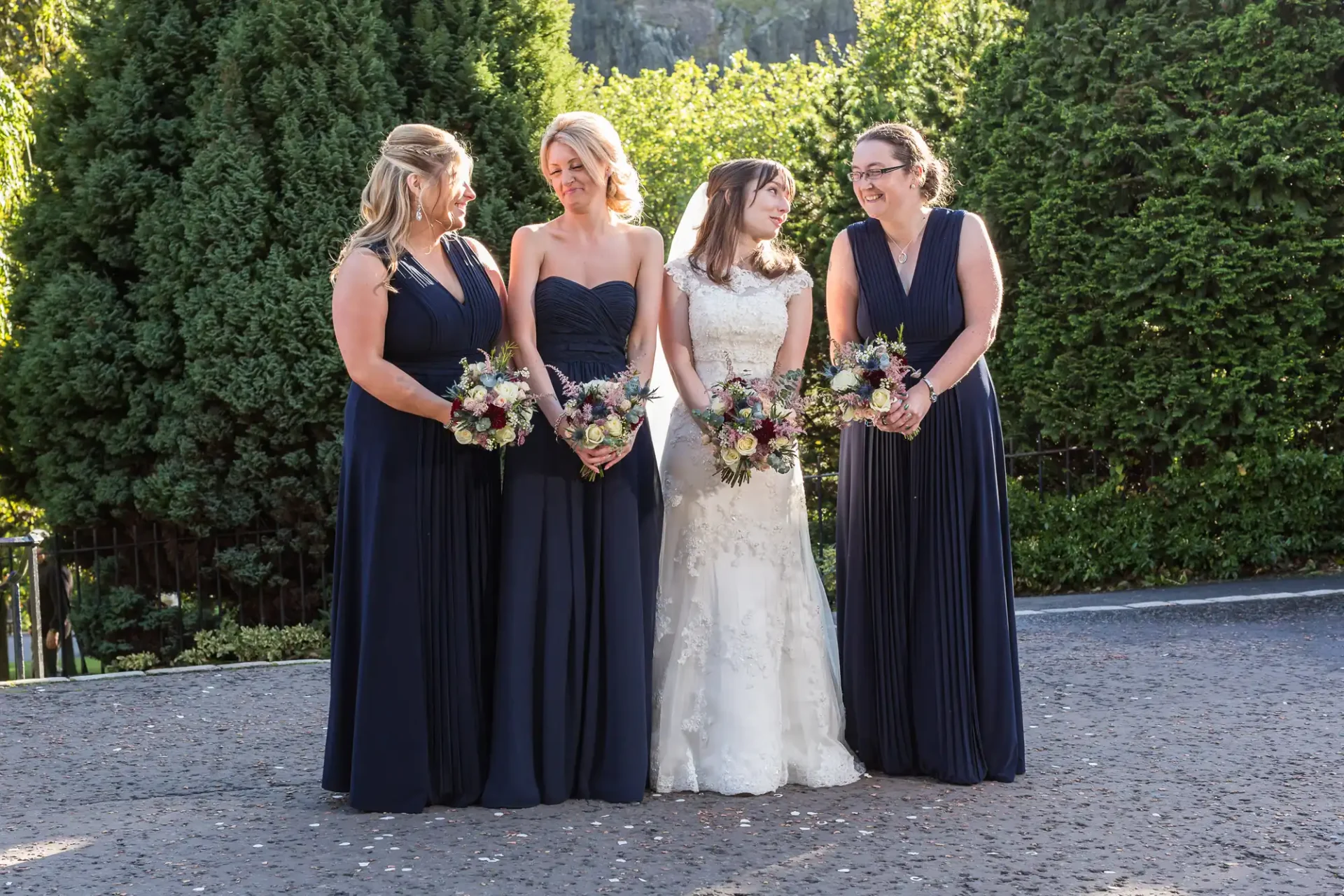 A bride in a white gown stands with three bridesmaids in navy dresses, holding bouquets, in a garden setting.