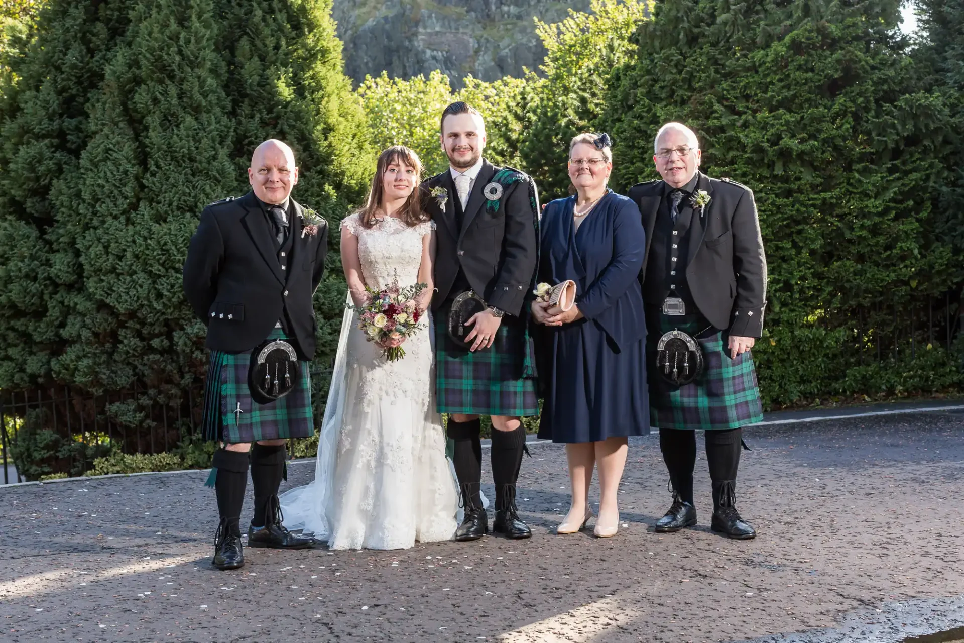 A wedding group with a bride, groom, and three others in traditional scottish attire, including kilts, posed outdoors with a backdrop of tall trees.