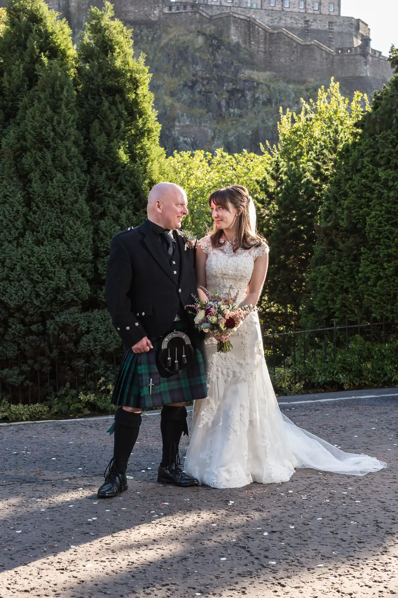 A bride in a white dress and a groom in a kilt stand smiling at each other, holding hands in a sunny park with a castle in the background.
