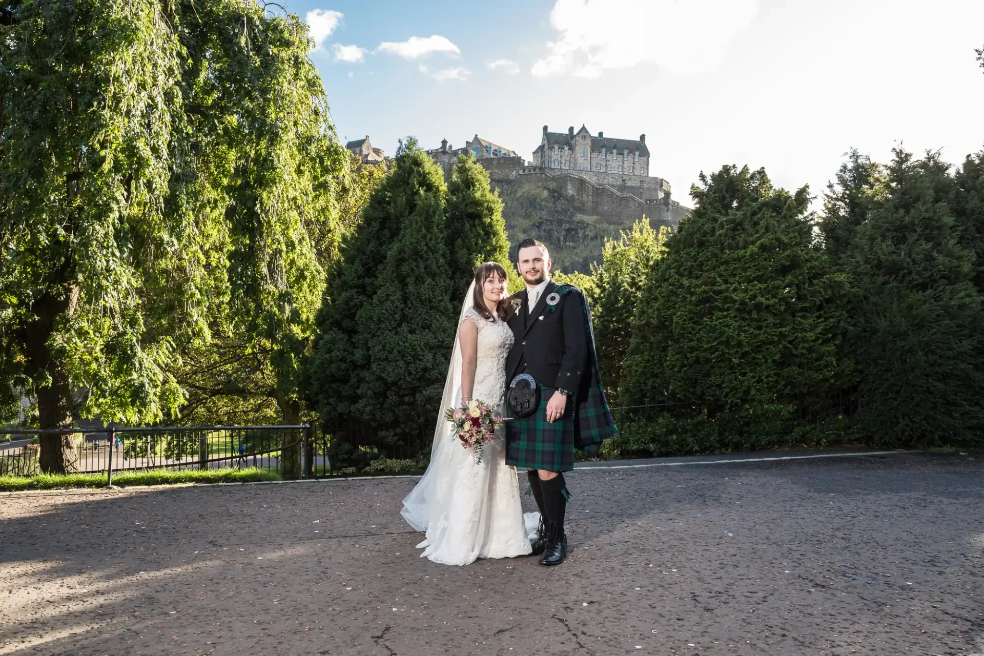A bride in a white dress and a groom in a kilt stand in a park with edinburgh castle in the background.