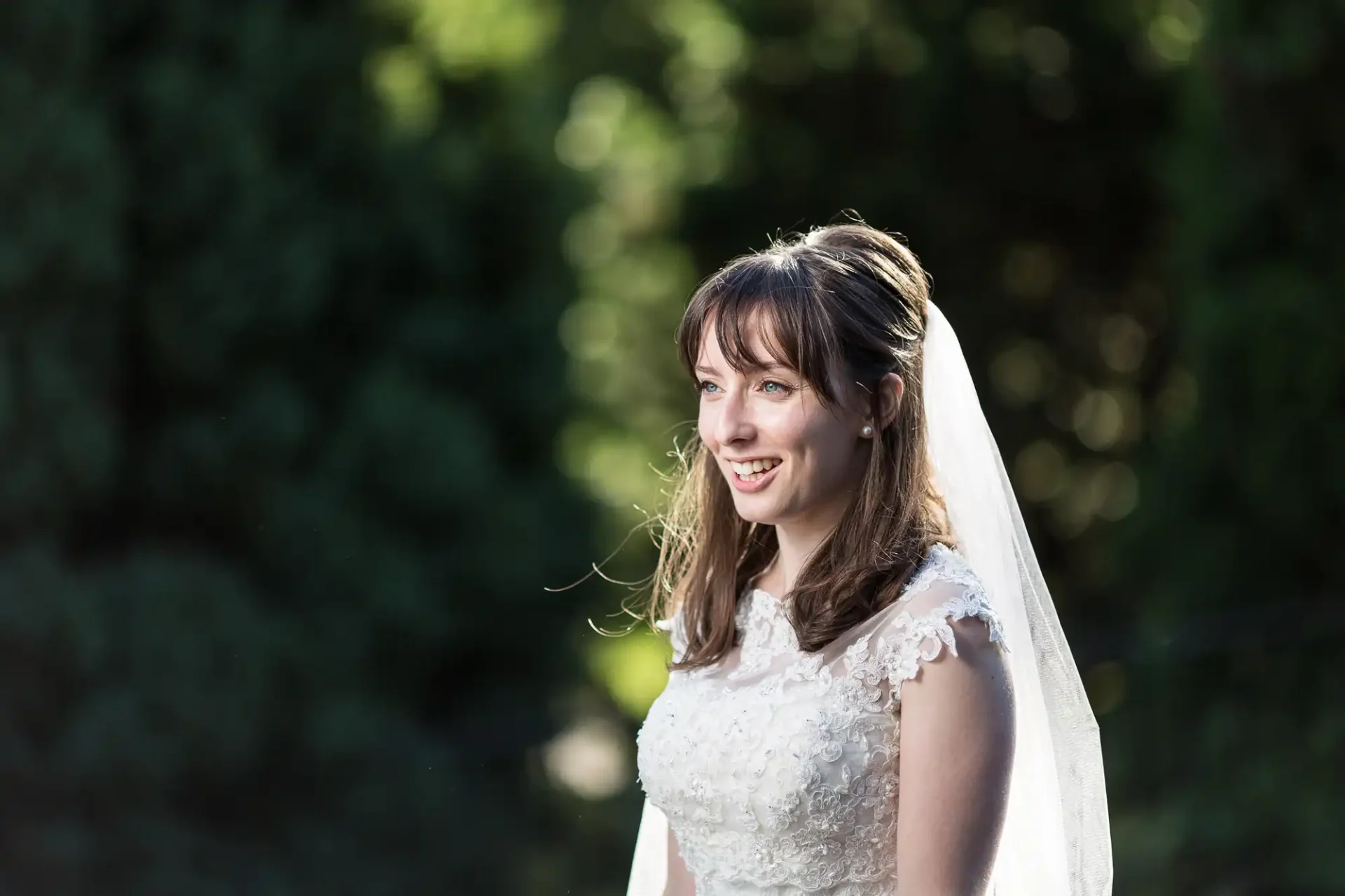 A smiling bride in a lace gown and veil, backlit by sunlight, with a blurred greenery background.