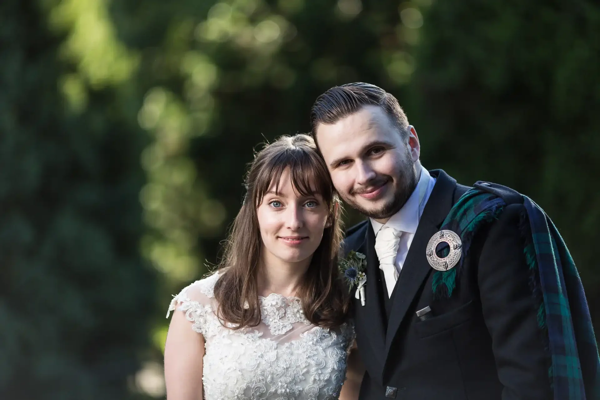 A bride and groom smiling outdoors, with the groom wearing a tartan kilt and the bride in a lace dress.