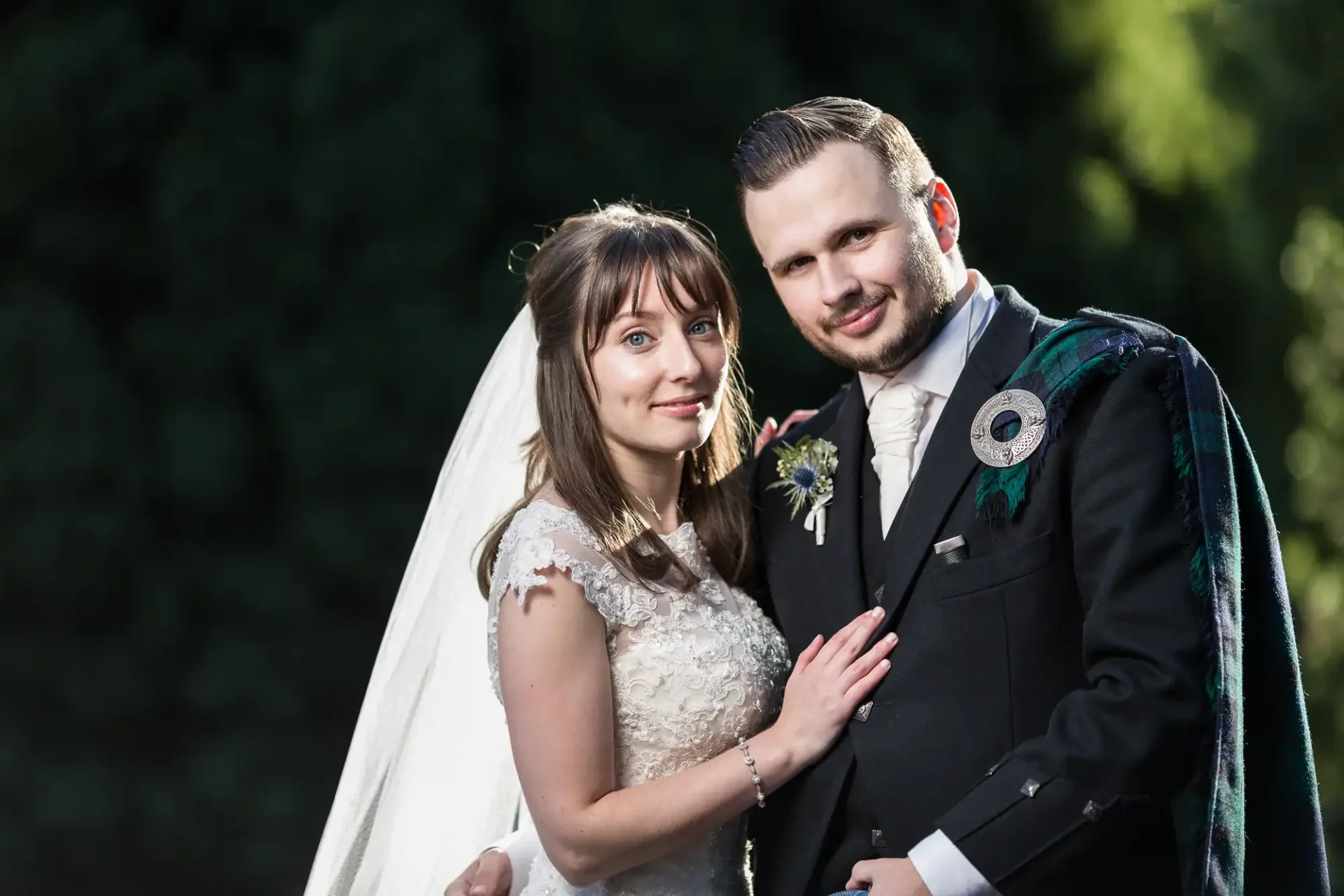 A bride in a white dress and a groom in a traditional kilt posing outdoors, with a green background.
