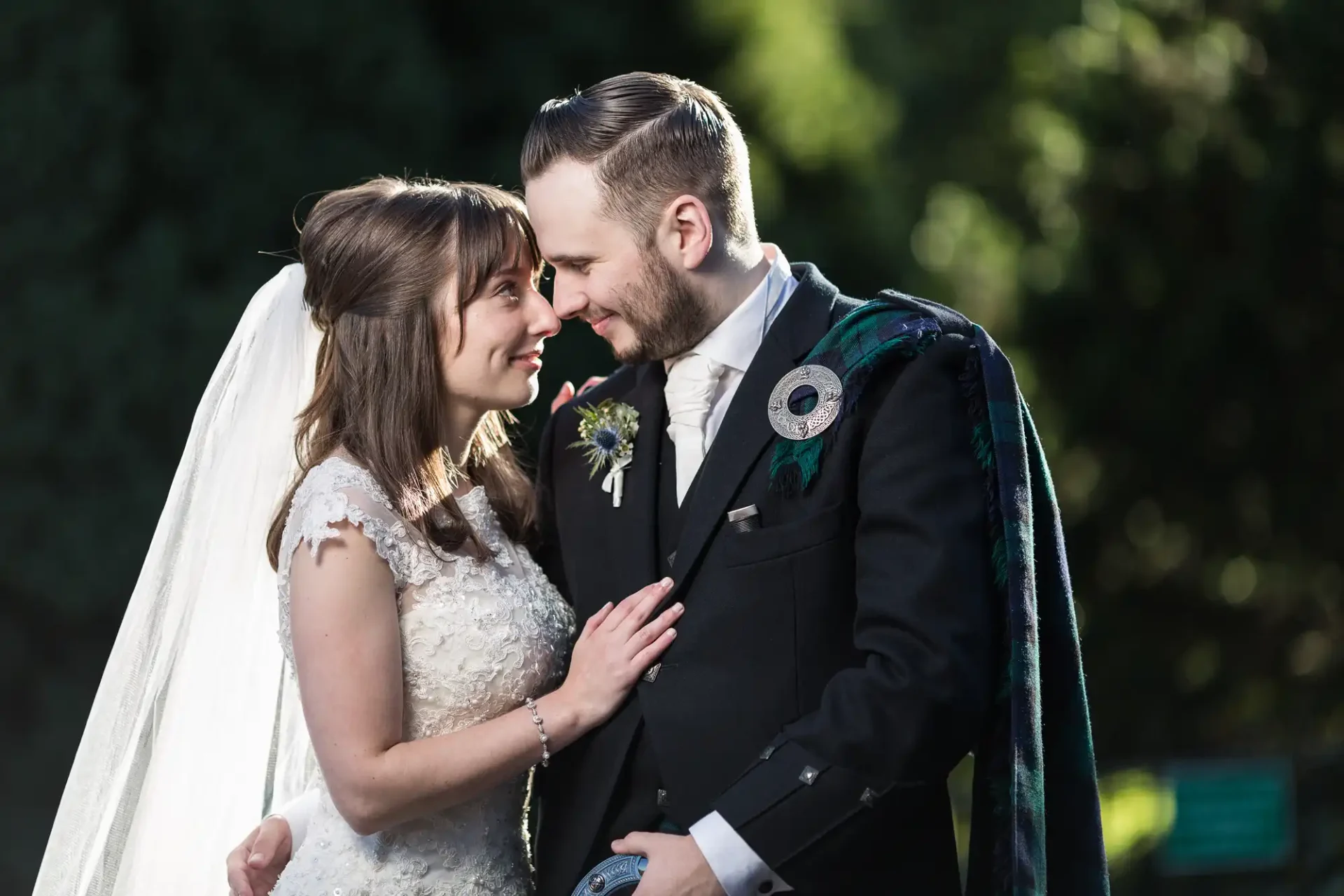 A bride and groom smiling at each other affectionately in a sunlit garden, dressed in wedding attire.