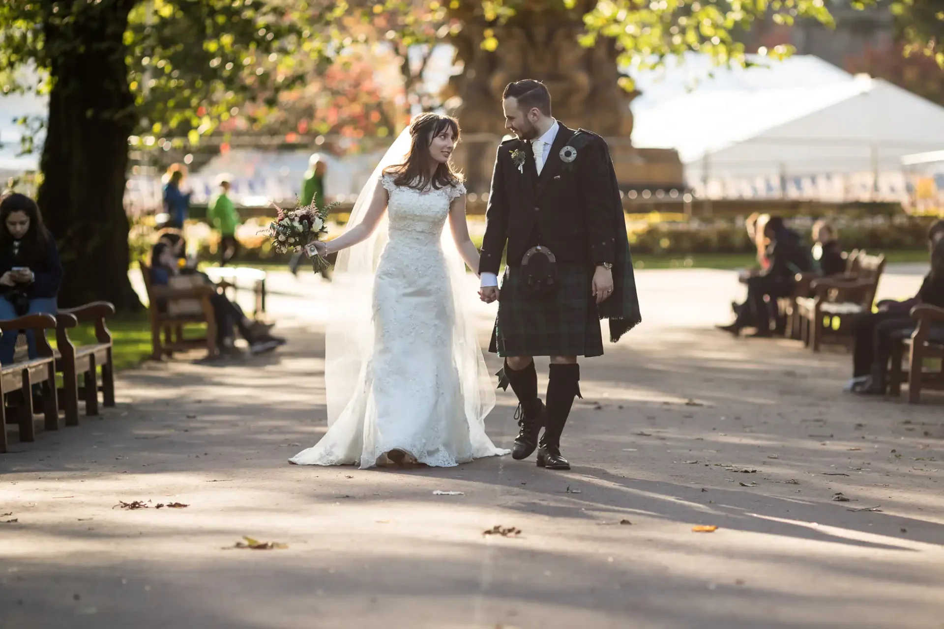 A bride in a white dress and a groom in a kilt walk hand in hand through a sunlit park with scattered leaves, smiling joyously.