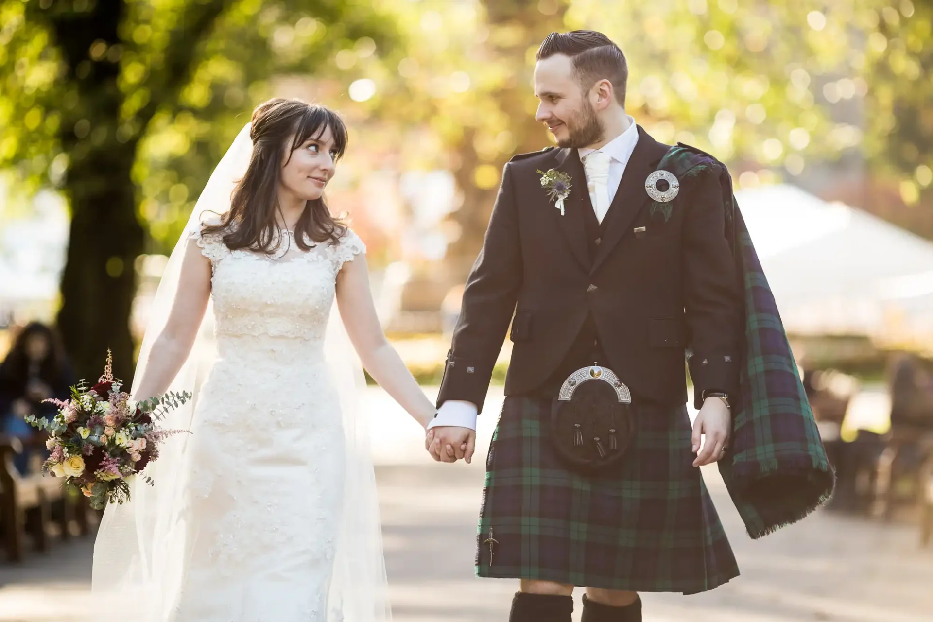A bride and groom holding hands, walking down a park path; the groom wears a kilt, and the bride is in a lace dress.