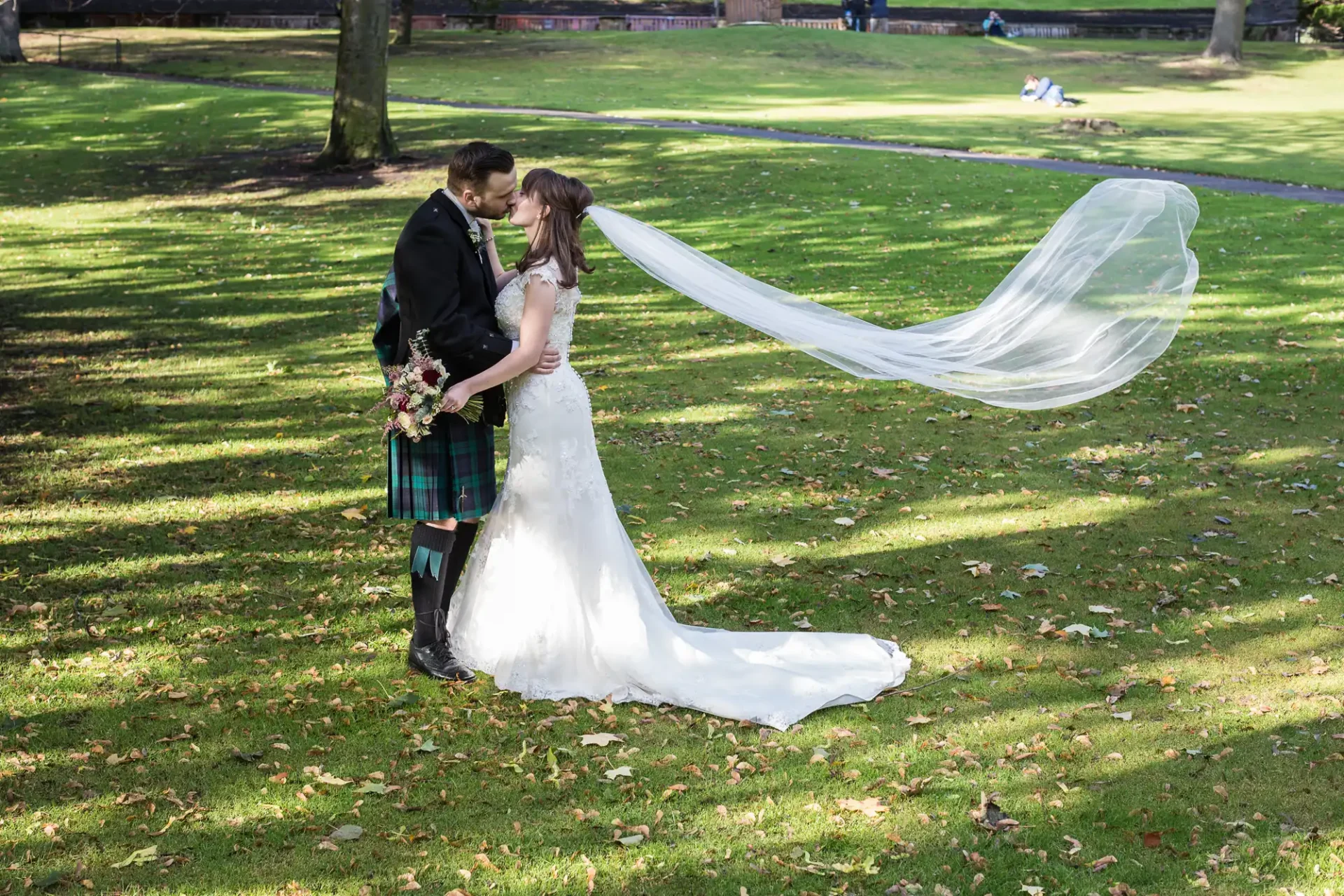 A bride and groom embracing in a park, the bride's long veil flowing in the breeze, with trees and grass around them.