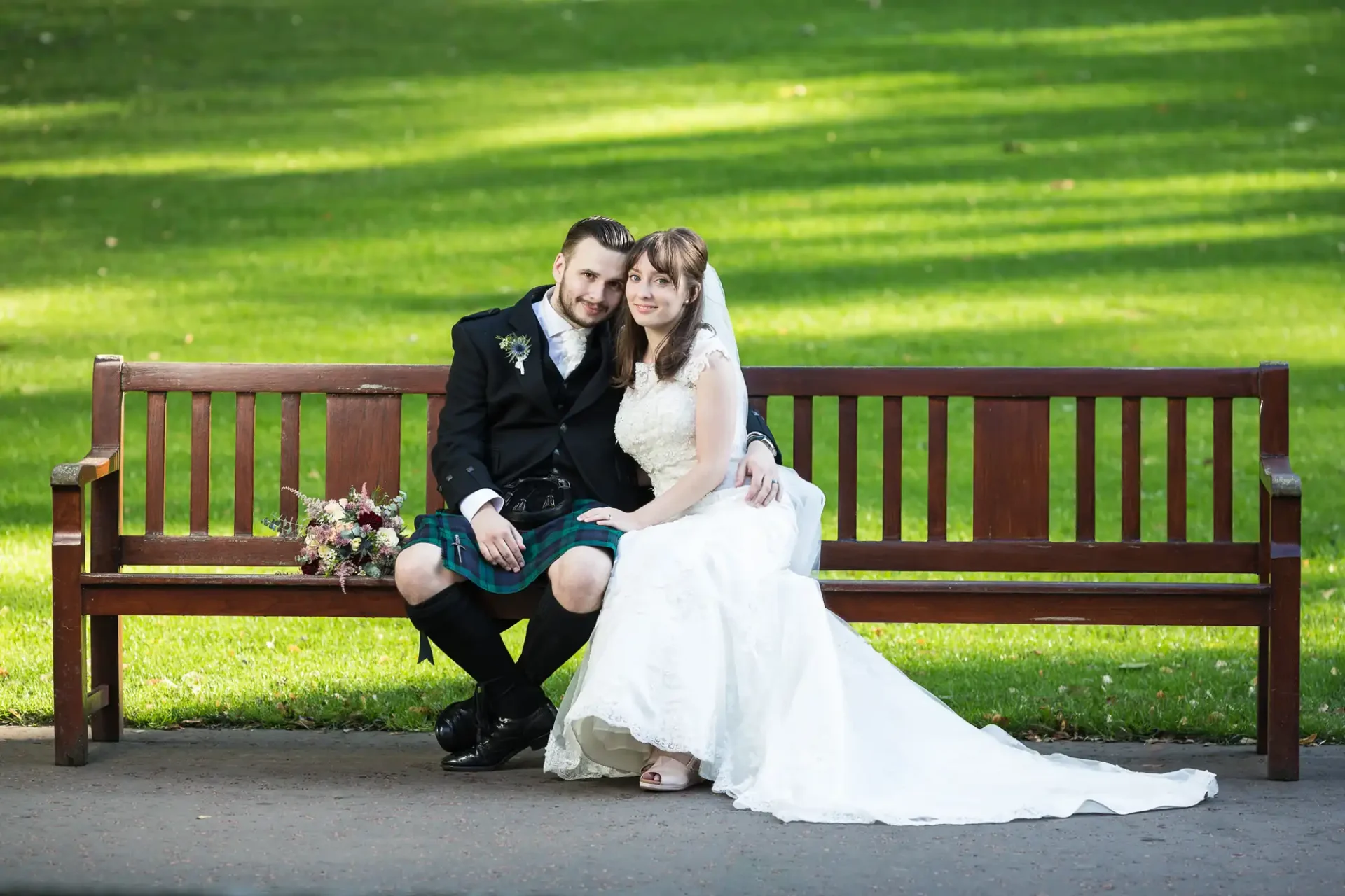 A newlywed couple in wedding attire sitting on a park bench, smiling at the camera, surrounded by green grass.
