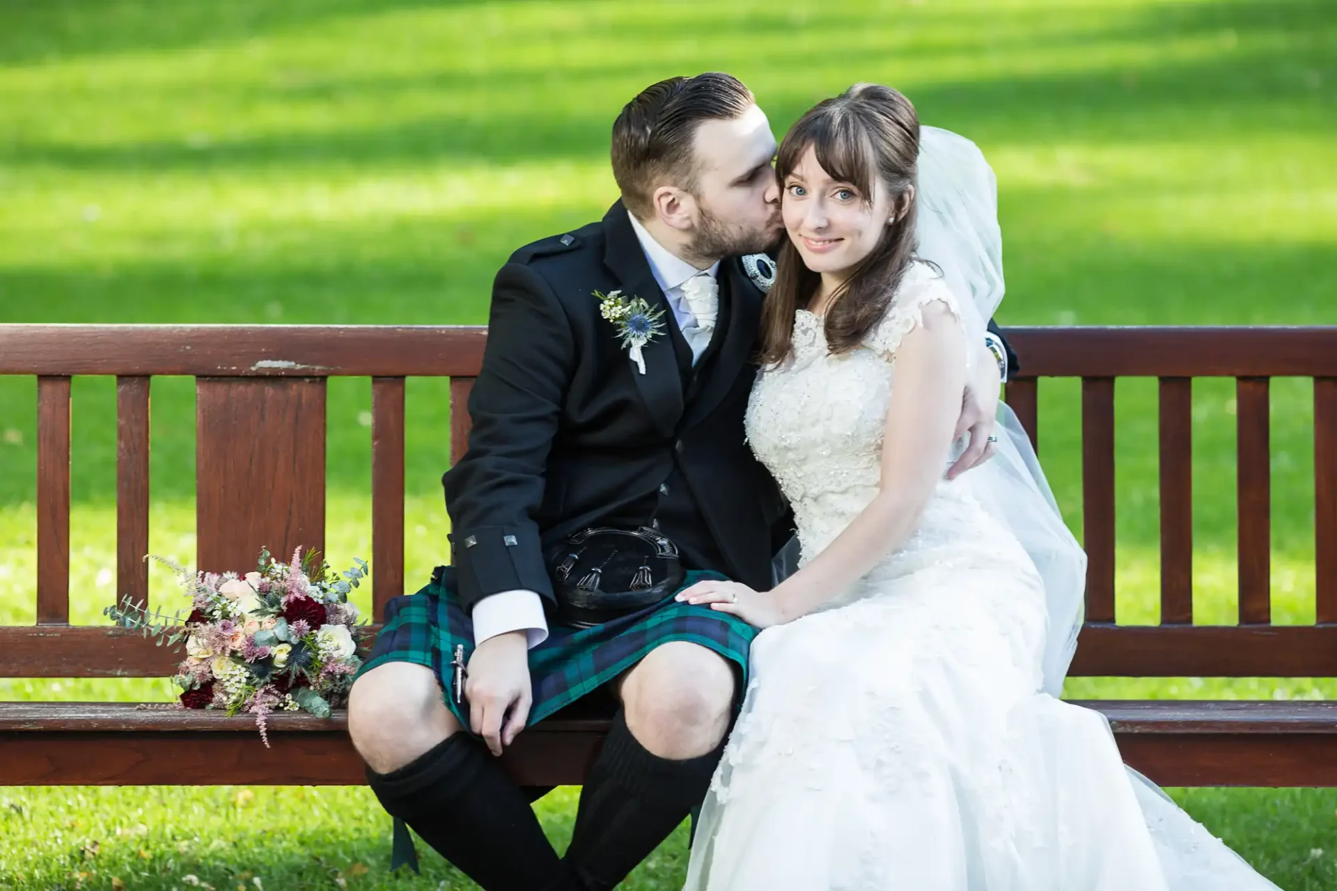 A groom in a kilt kisses a bride on the cheek as they sit on a park bench, both smiling, dressed in wedding attire.
