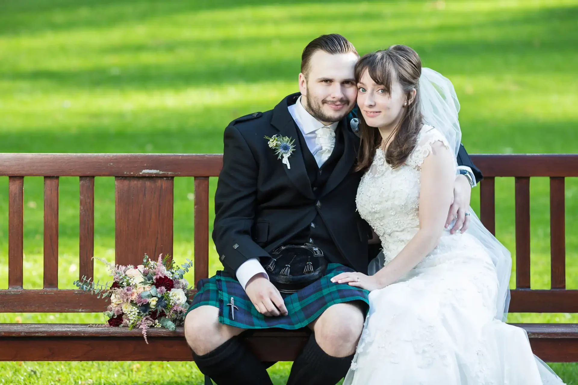 A bride and groom smiling and sitting closely on a park bench; the groom is wearing a kilt and the bride a white dress with a bouquet beside them.