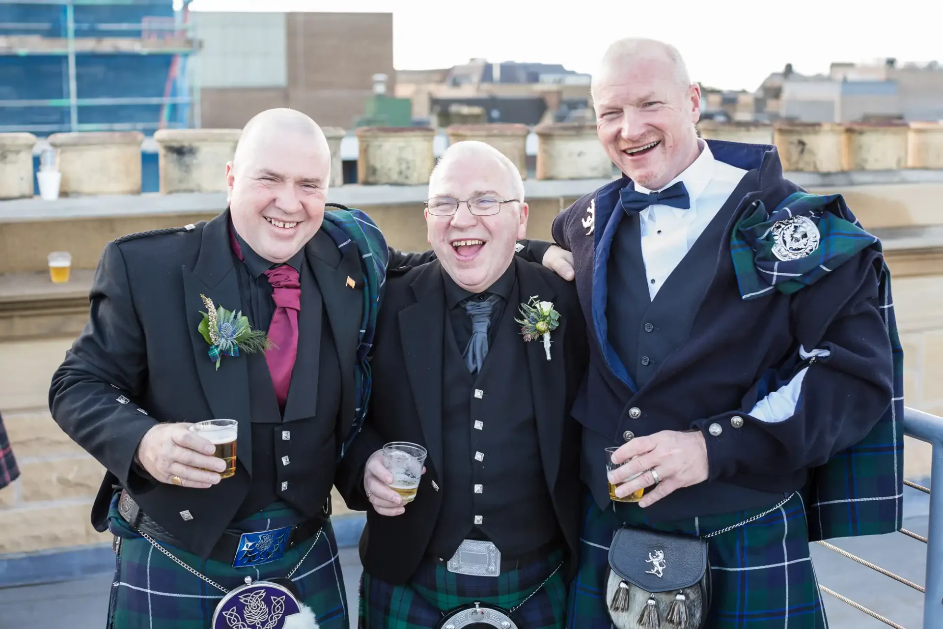 Three smiling men in traditional scottish attire holding drinks at an outdoor event.