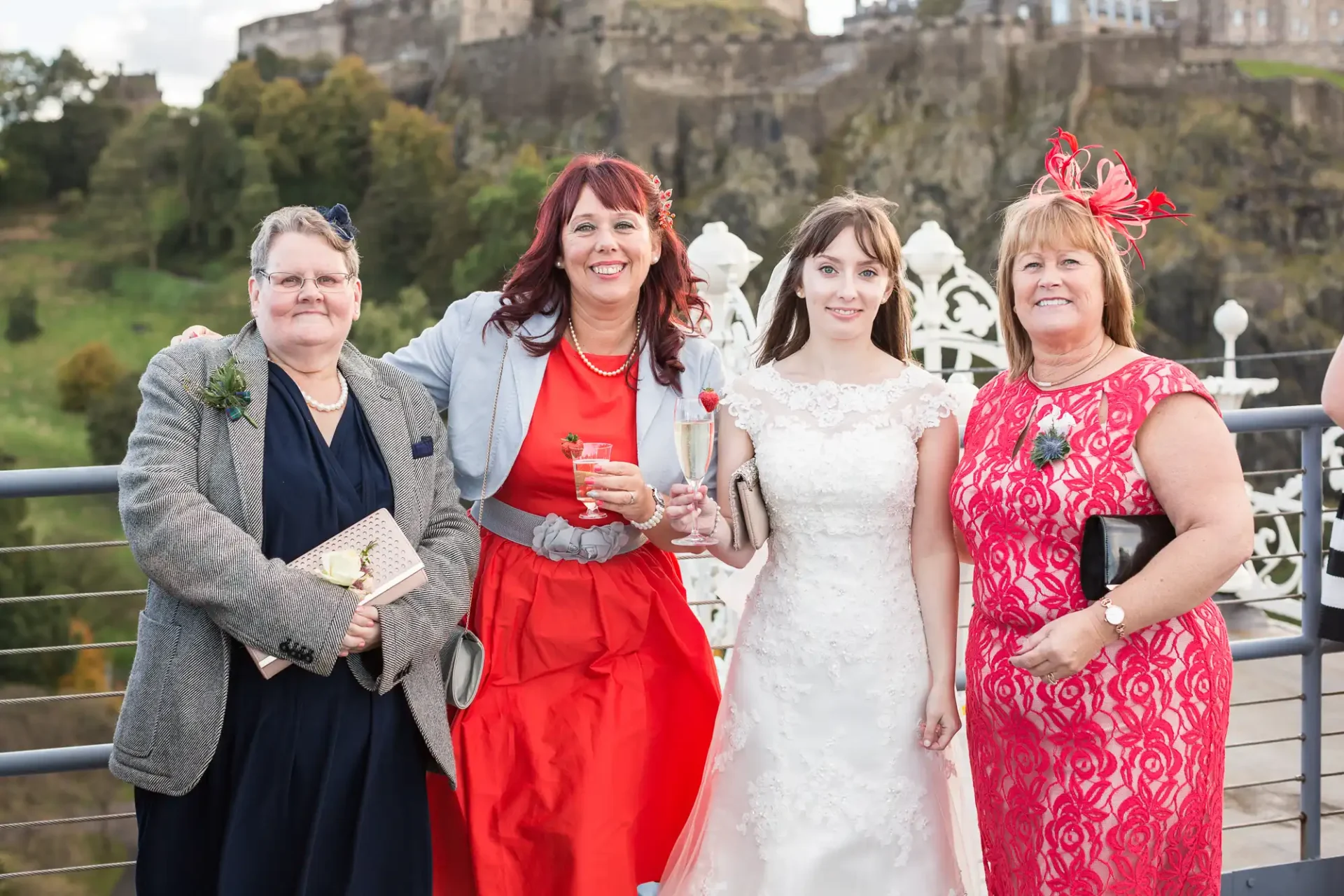 Four women in formal attire pose for a photo outdoors, with a castle in the background. the woman in a white bridal gown stands center, flanked by three others wearing colorful dresses.