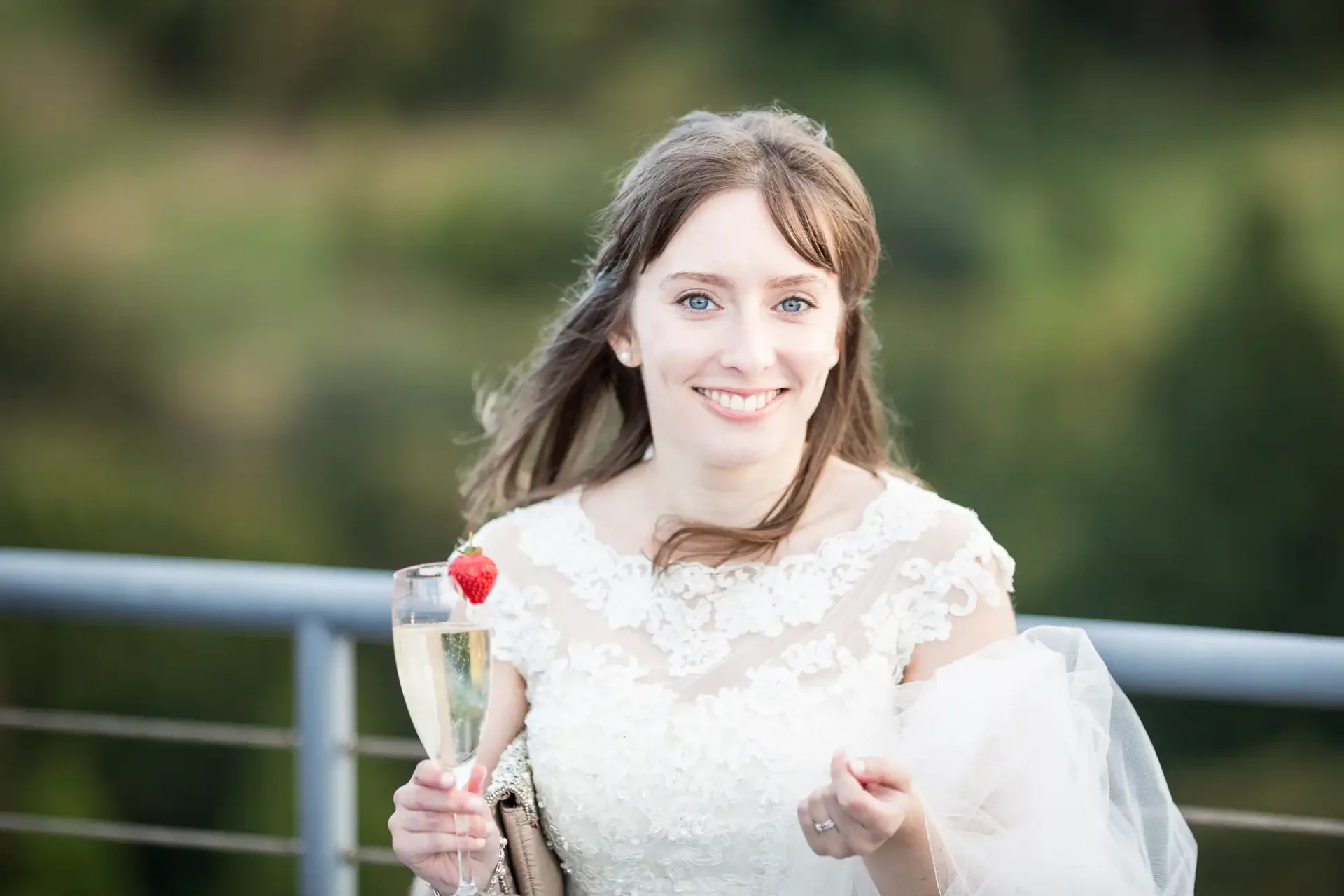 A smiling bride in a lace dress holding a champagne glass with a strawberry, standing outdoors with a blurred green background.