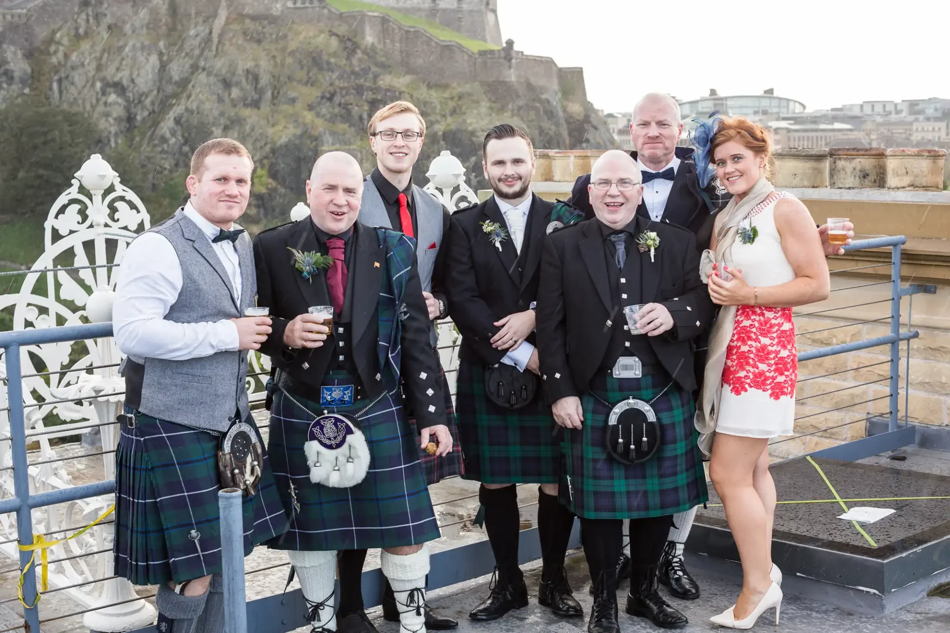 Group of seven people, some in kilts, holding drinks and smiling on a terrace with a cityscape background.