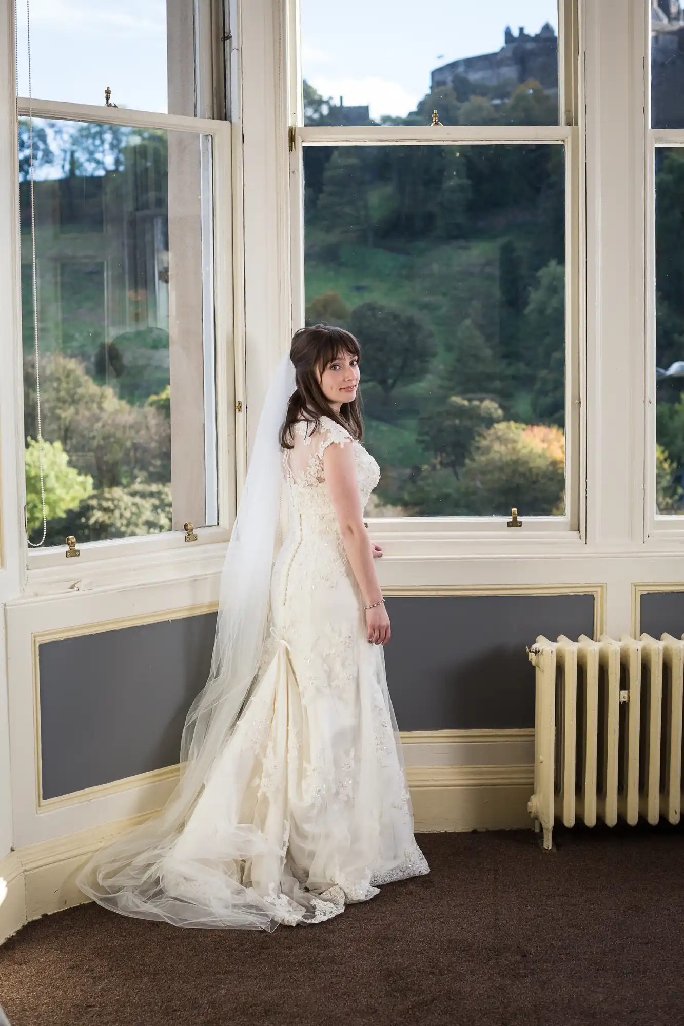 A bride in an elegant white dress stands by a window overlooking green hills, looking over her shoulder with a gentle smile.