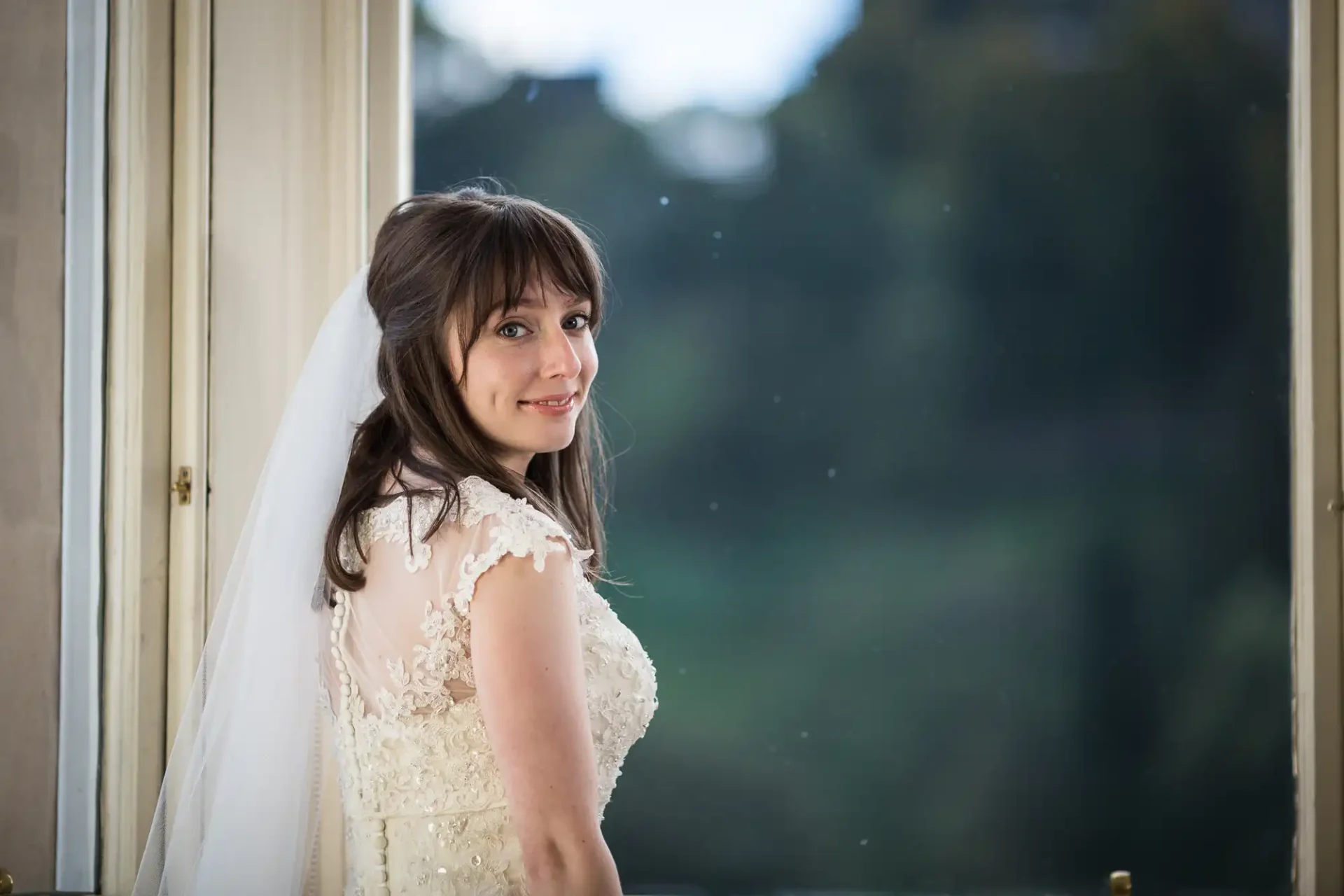 A bride in a lace wedding dress smiling over her shoulder near an open door, with a blurred green background.