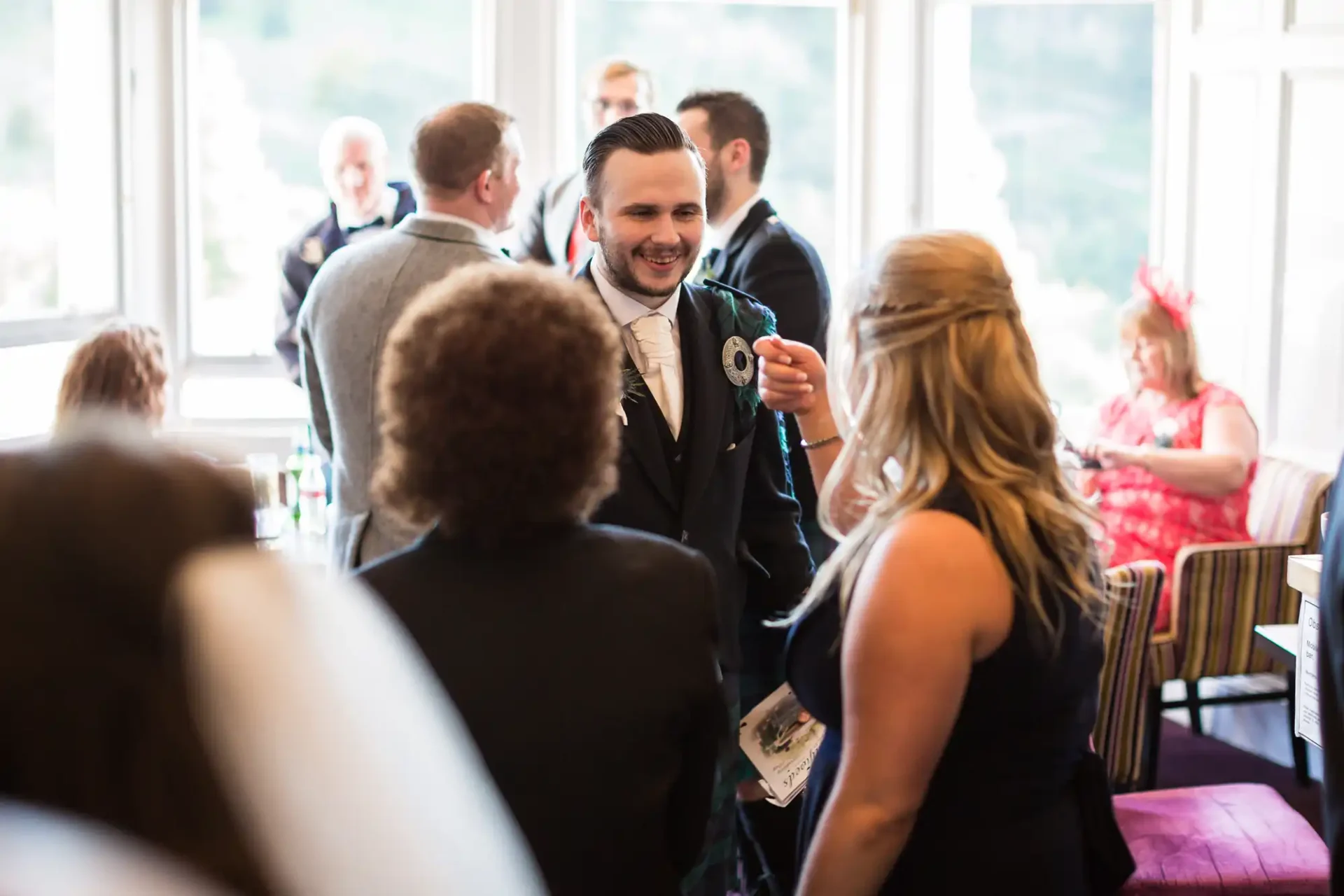 A man in a suit with a boutonniere smiles during a conversation at a lively indoor event with guests in the background. .