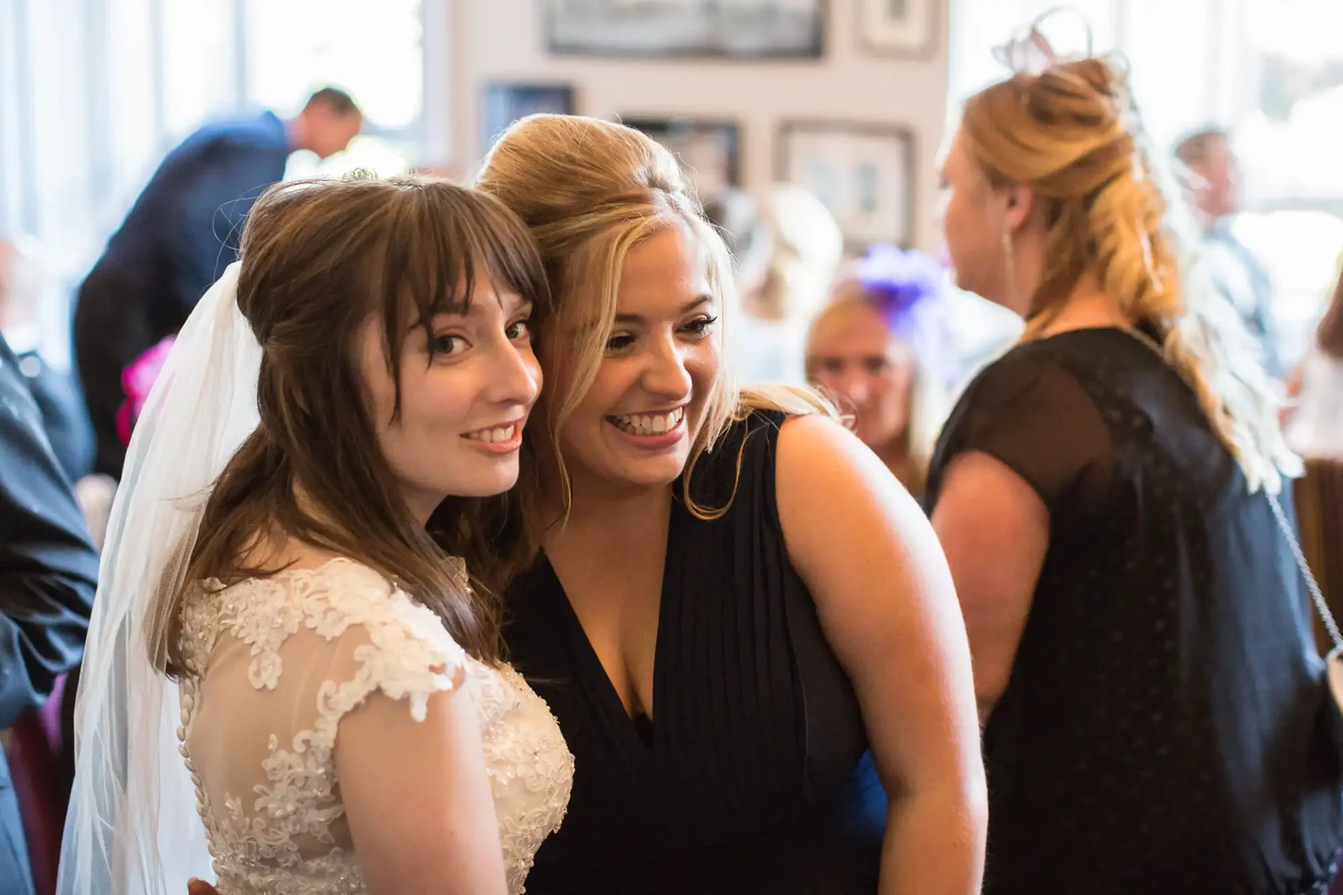 A bride in a white dress and a woman in a black dress smiling together at a wedding reception.
