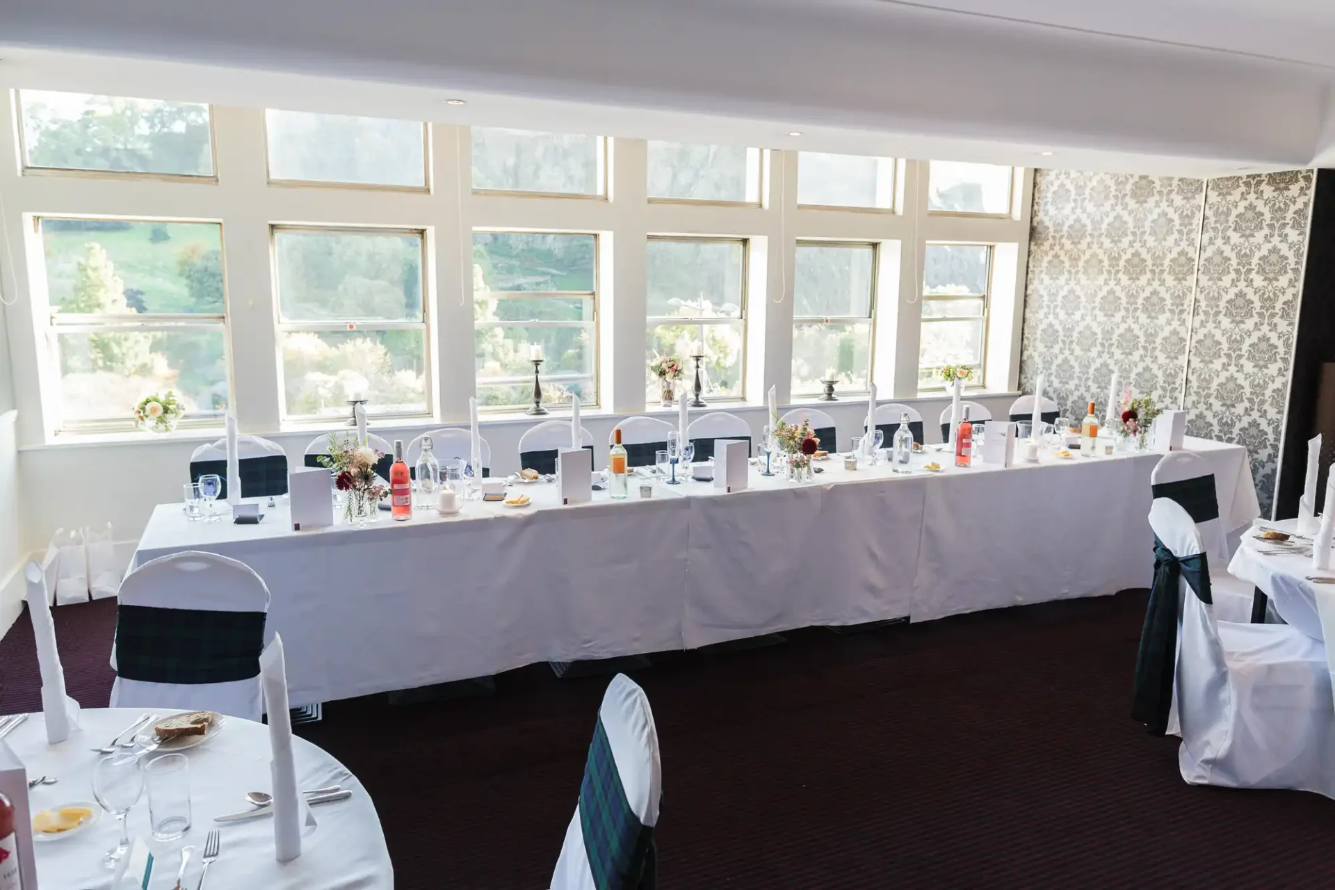 Elegant dining room setup for an event, with a long table covered in white linen, chairs in black covers, and various table settings under large windows with a scenic view.