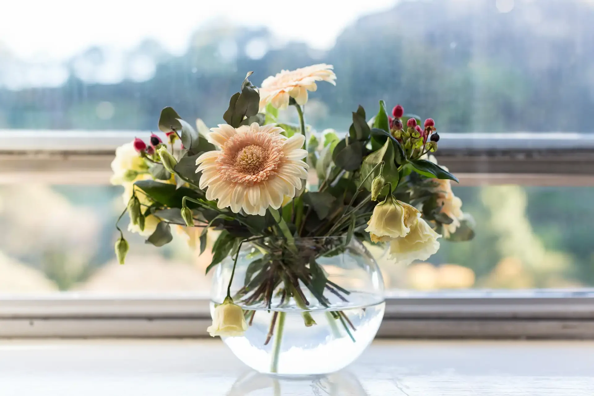 A bouquet of fresh flowers in a glass vase on a windowsill with a rainy landscape visible through the glass.