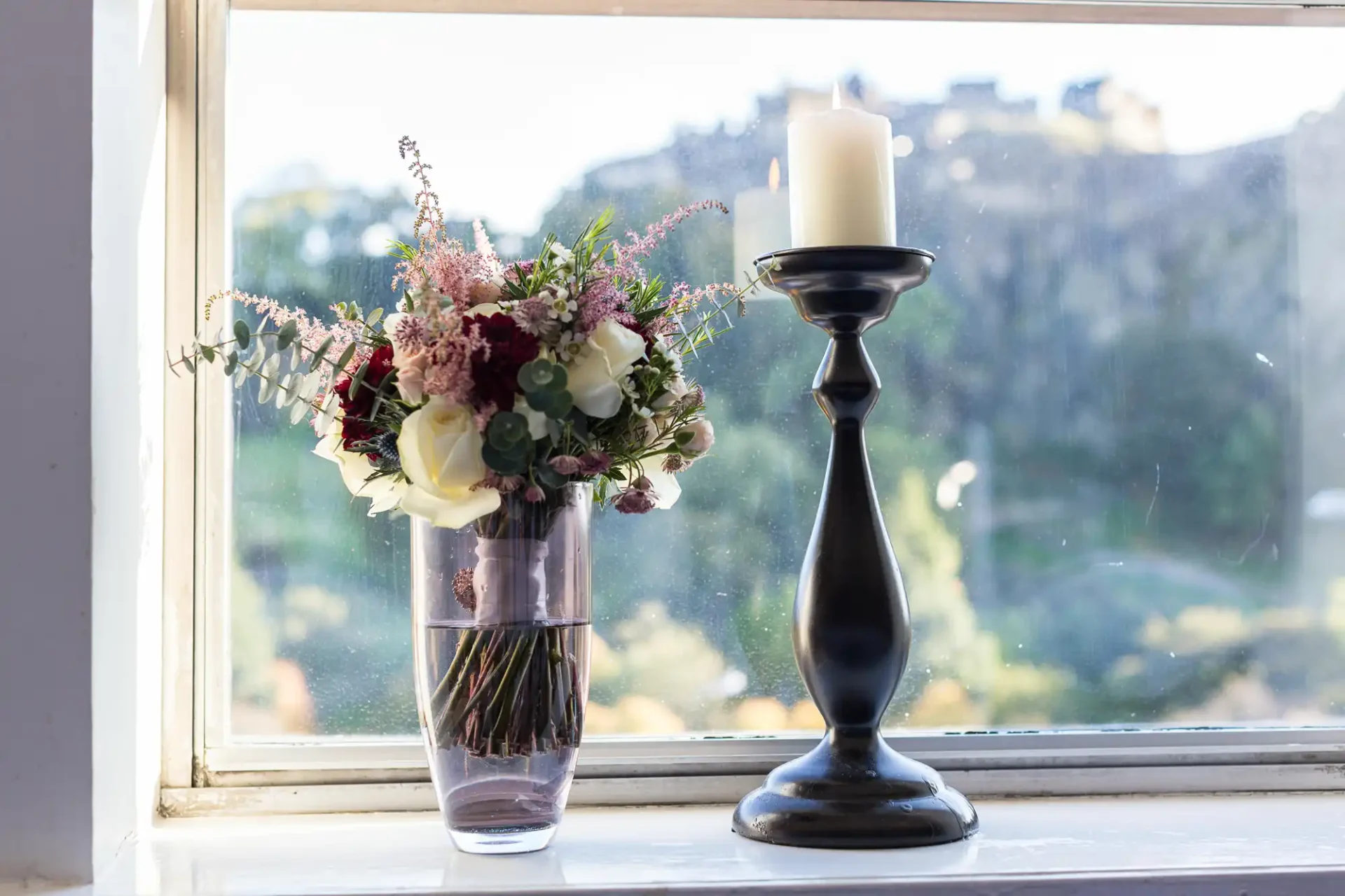 A bouquet of flowers and a lit candle on a tall black holder, placed on a windowsill with views of trees outside.
