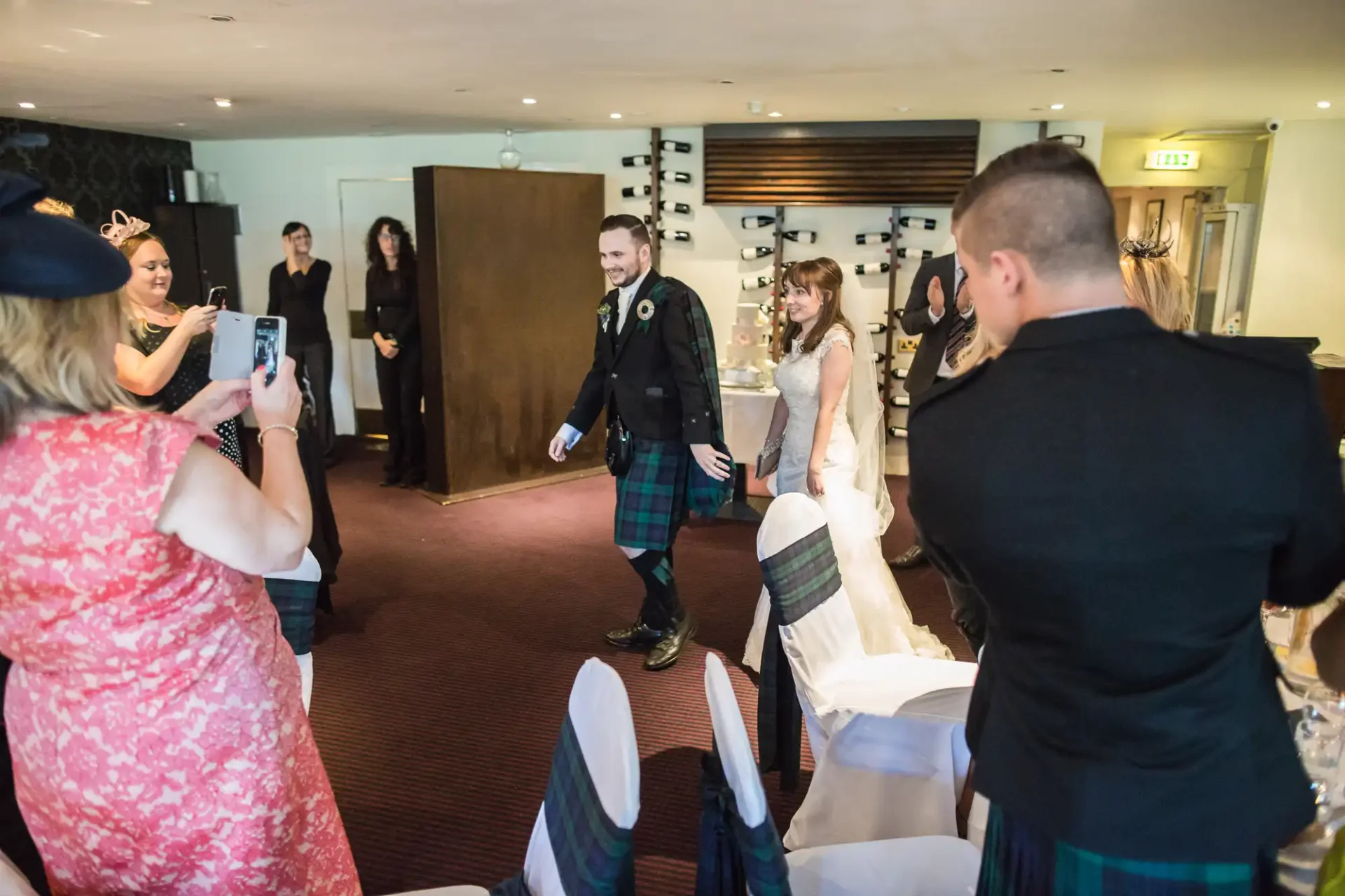 A bride and groom walking hand in hand through a room of guests, with the groom wearing a traditional kilt.