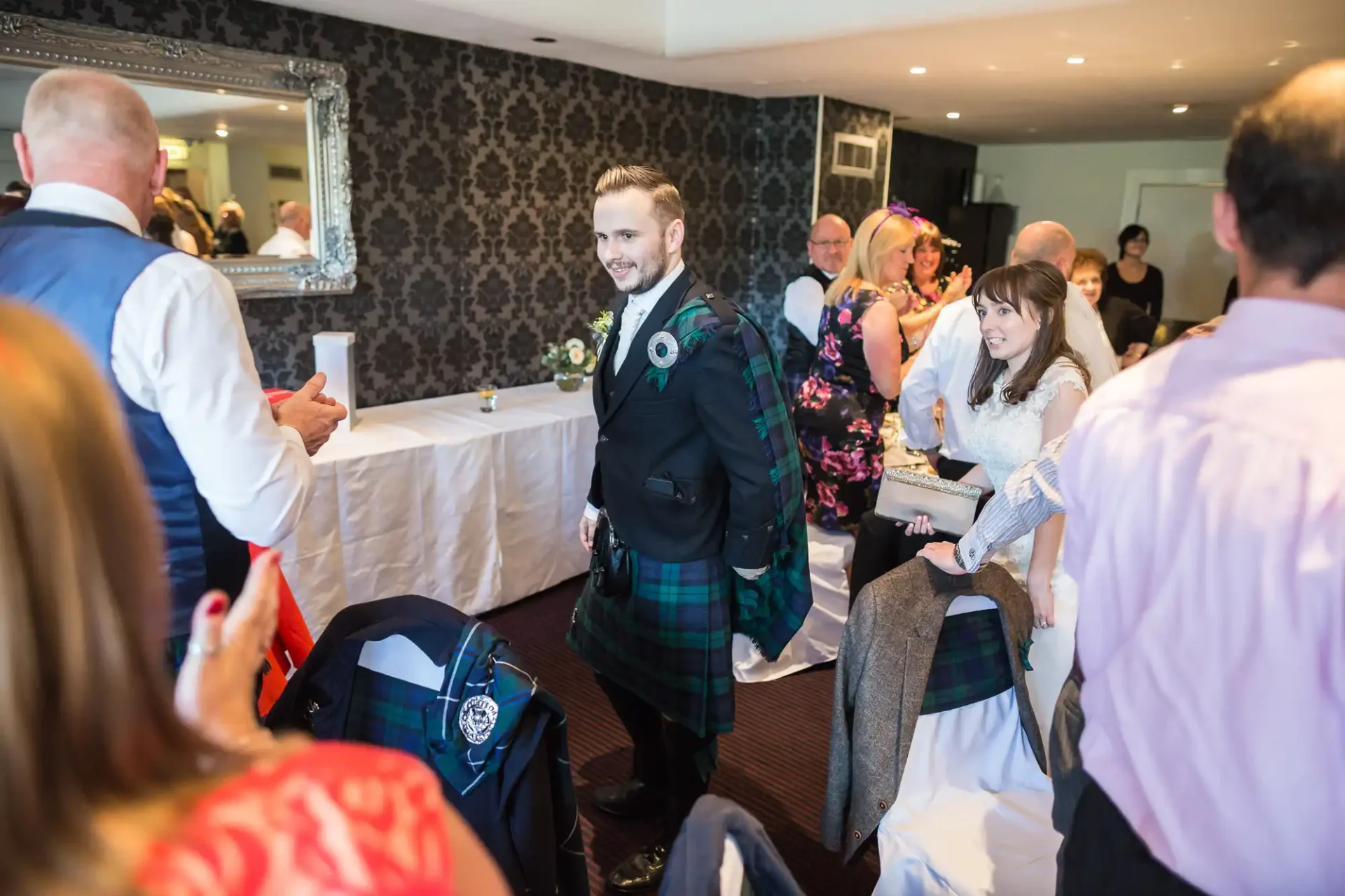 A groom in a tartan kilt smiling as he walks through a crowd of guests at a wedding reception, with attendees clapping and smiling.