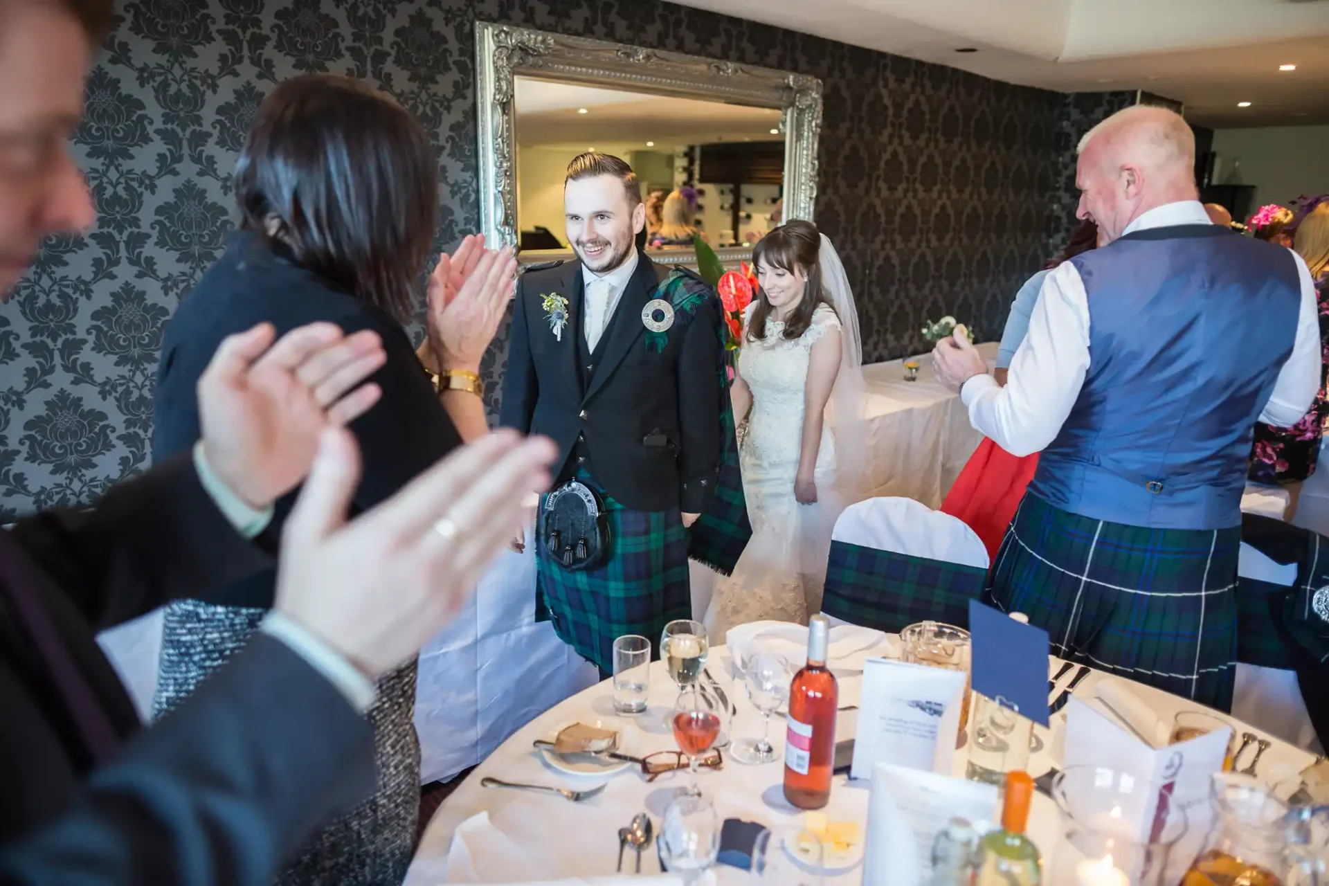 A bride and groom walking through a clapping crowd at a wedding reception, with guests wearing scottish kilts.