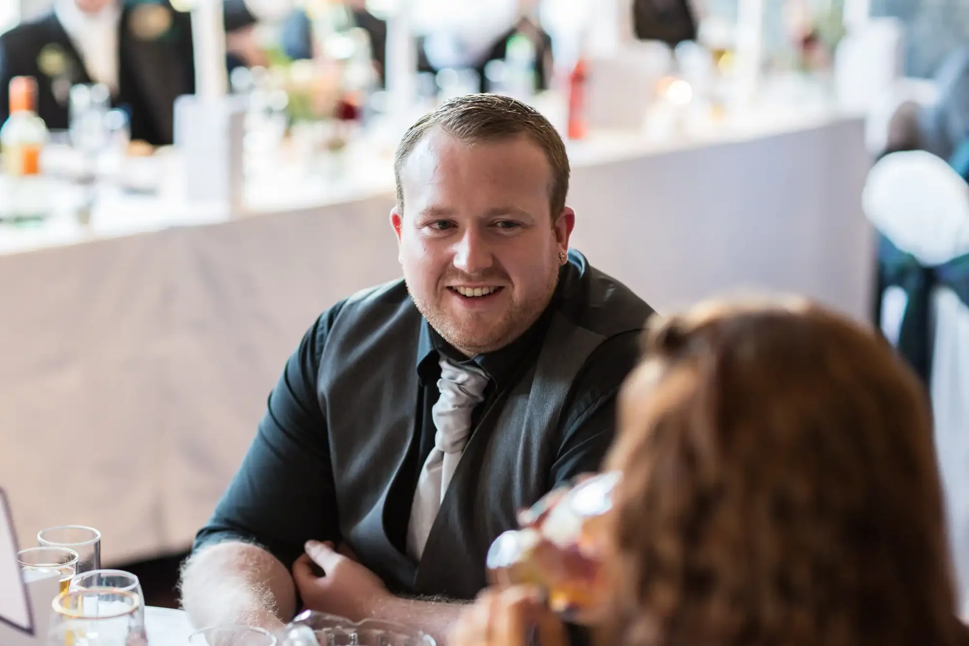 A man in a formal suit smiling at a table during an event, with blurred people and glasses in the background.