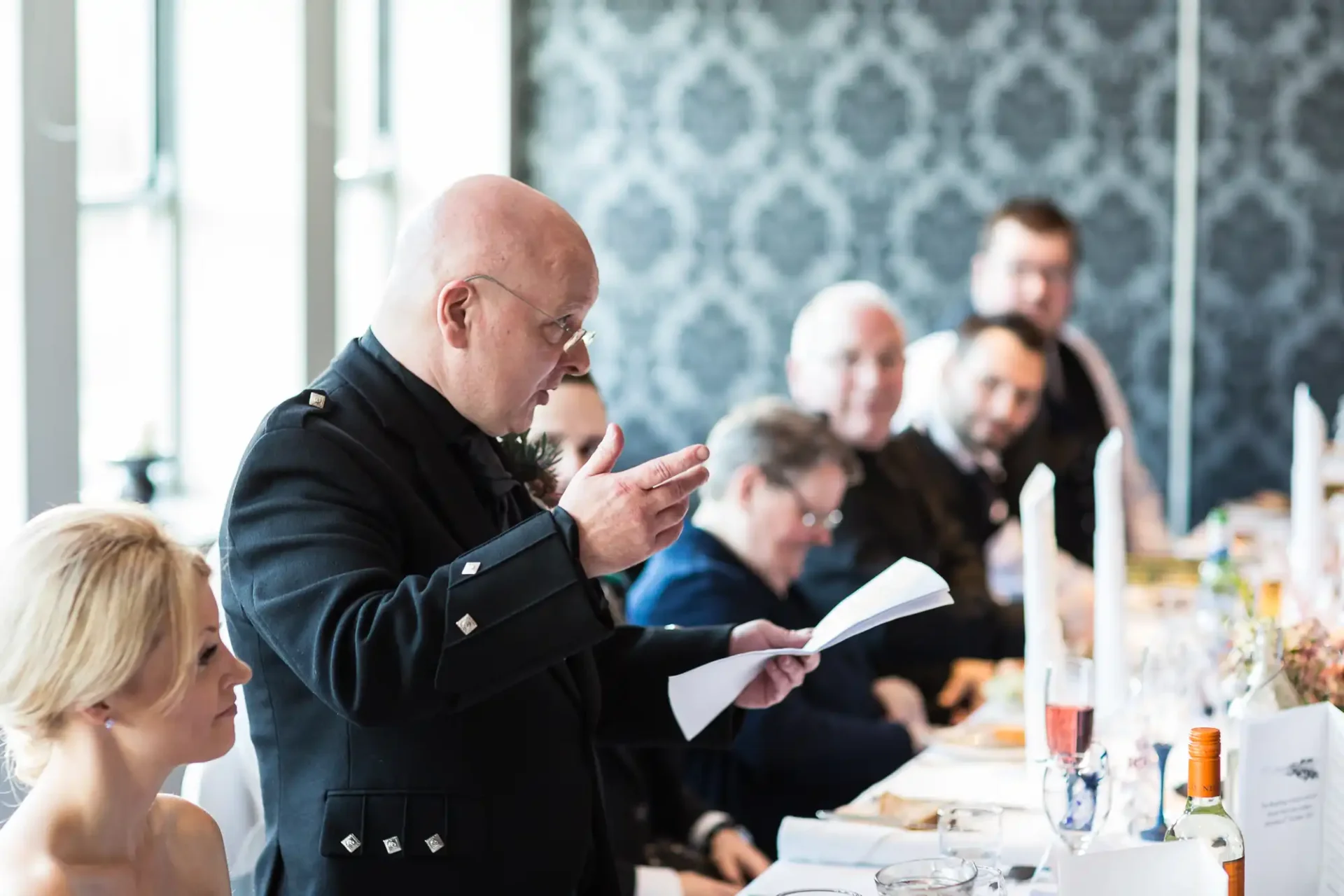 A bald man in a formal uniform speaking and gesturing with a paper at a dinner table, with a focus on seated guests listening.