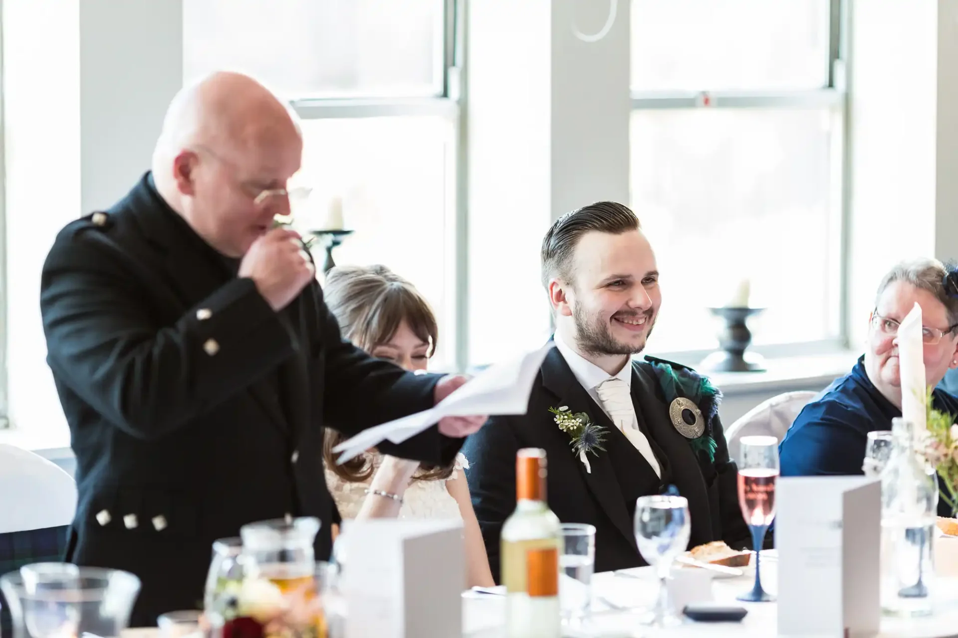Man in a suit smiling at a wedding table while another man laughs and covers his mouth with a napkin.