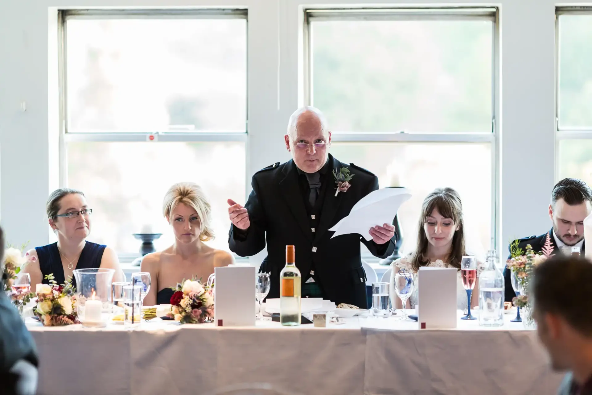 An elderly man reads from a paper at a wedding reception table, with guests seated around him looking attentive.