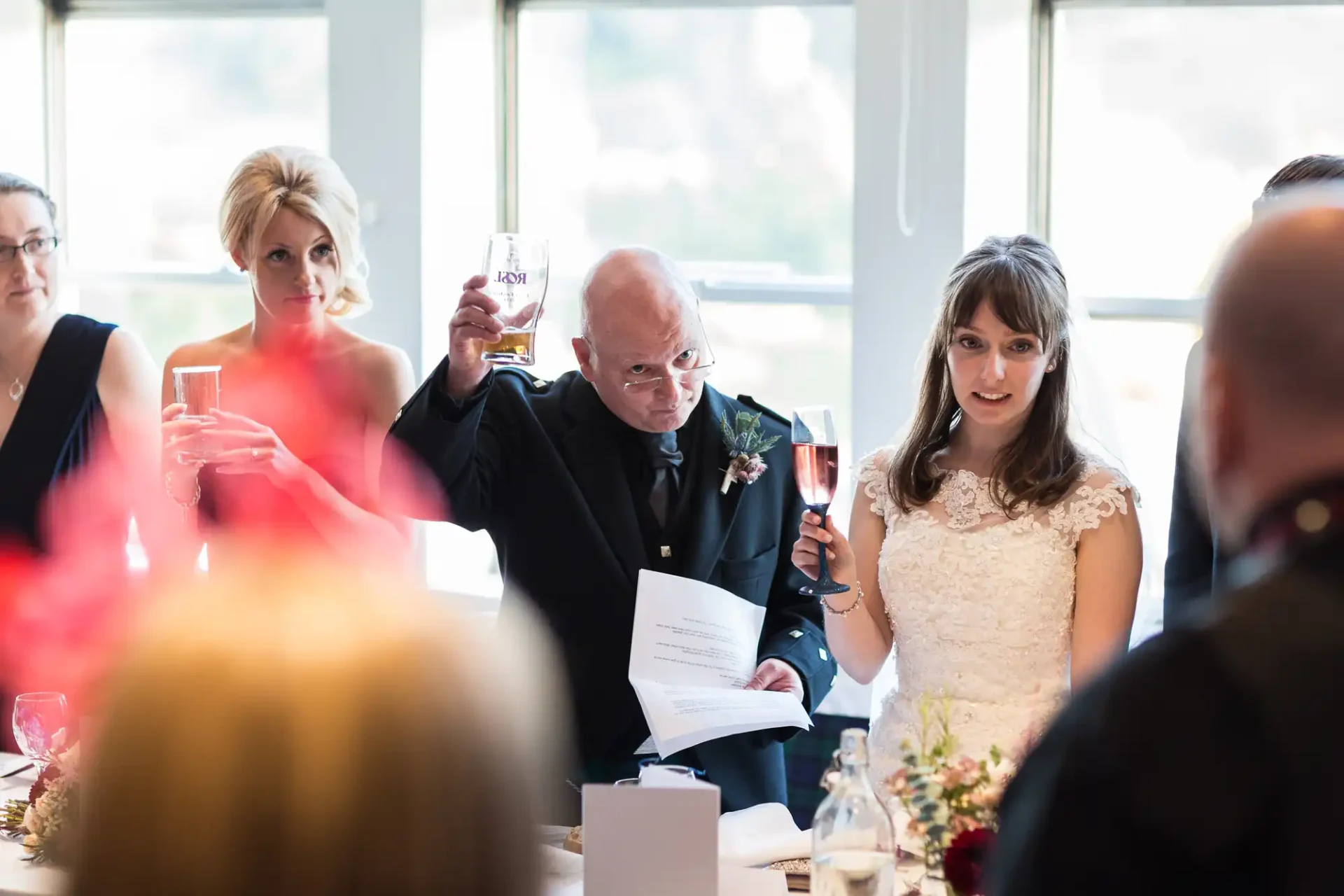 A bride and an older man raise their glasses for a toast at a wedding reception, observed by seated guests.