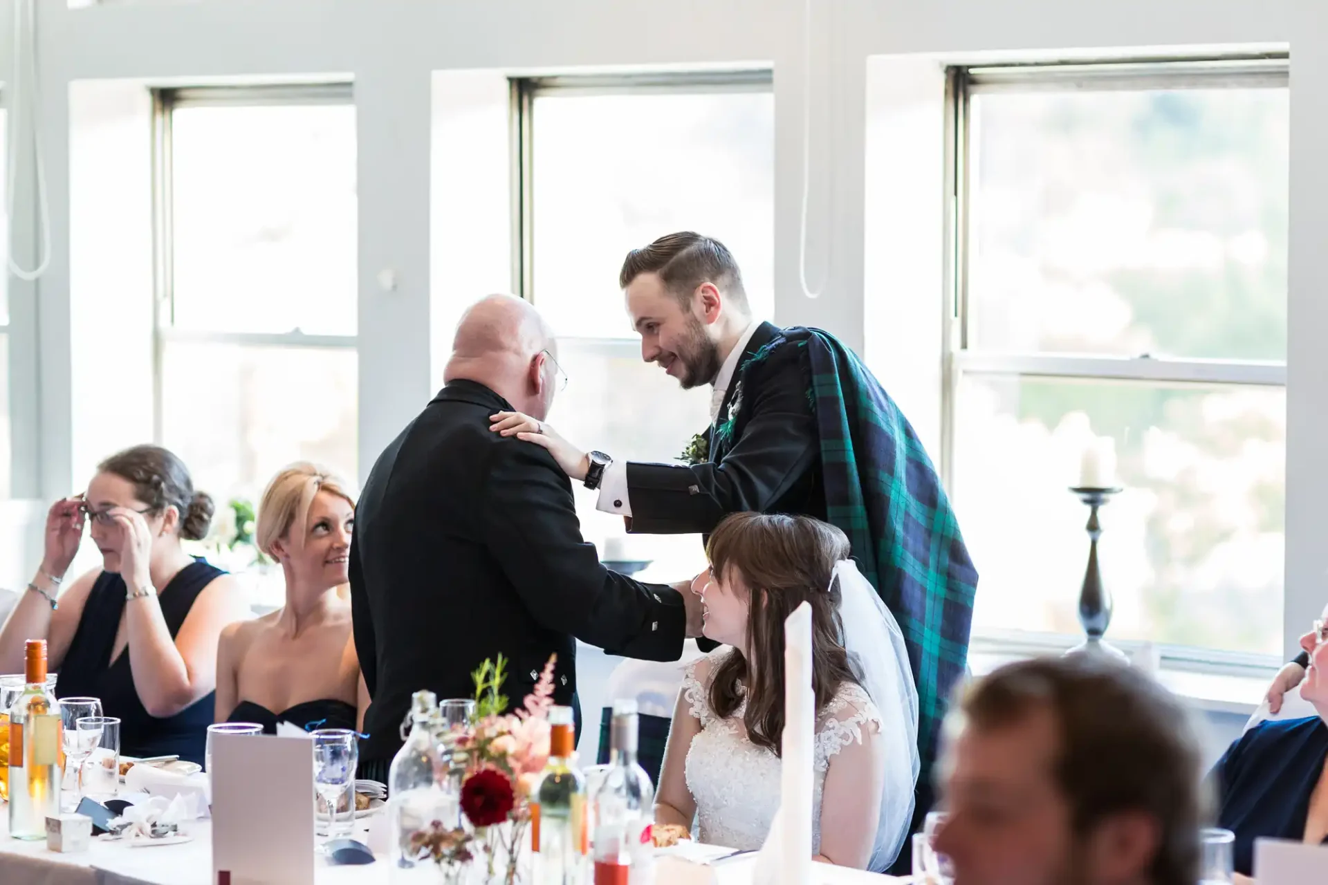 A groom in a kilt embraces an older man at a wedding reception, with guests looking on and smiling.