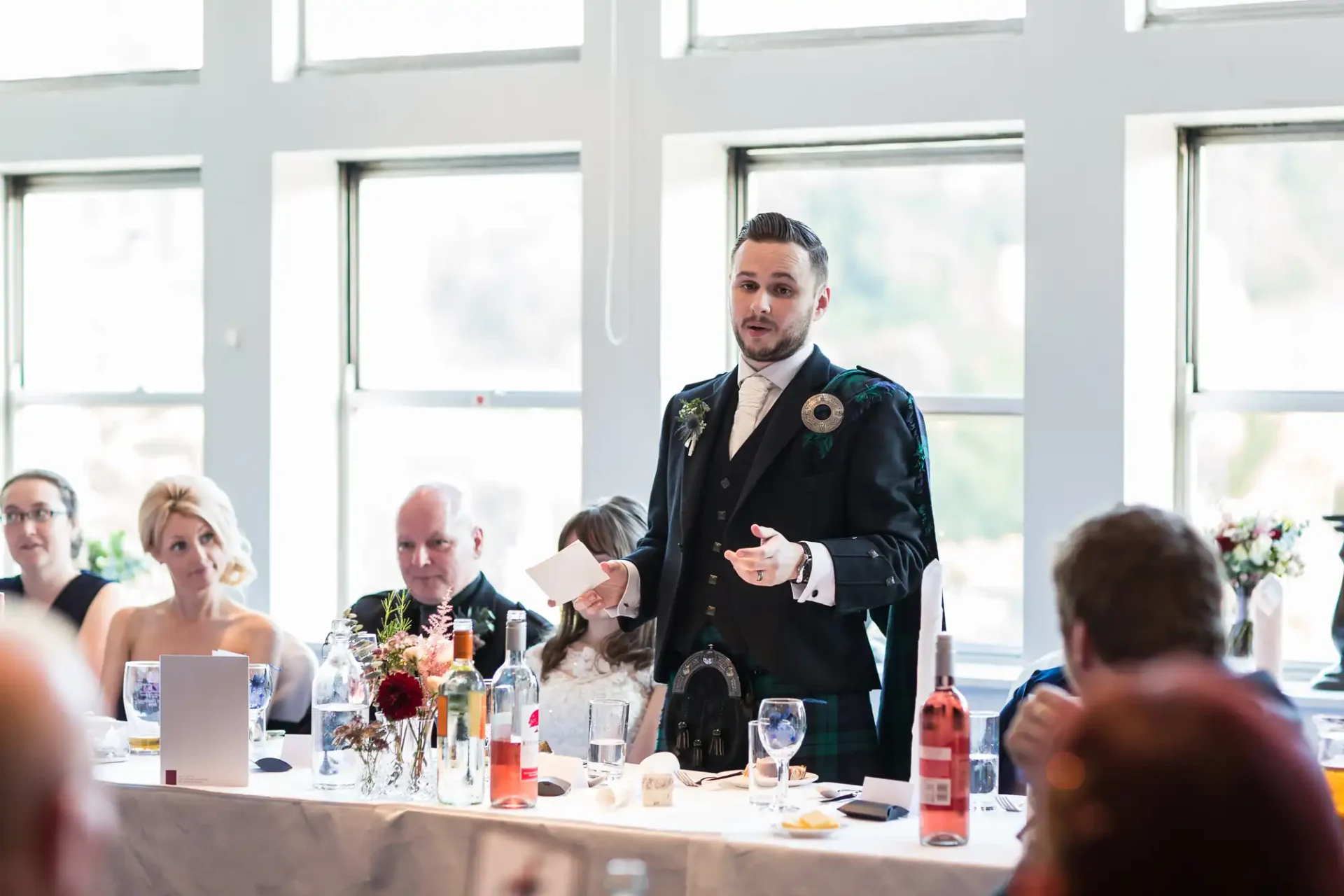 A man in a suit giving a speech at a wedding reception, holding a microphone in one hand and notes in the other, with guests listening around tables.