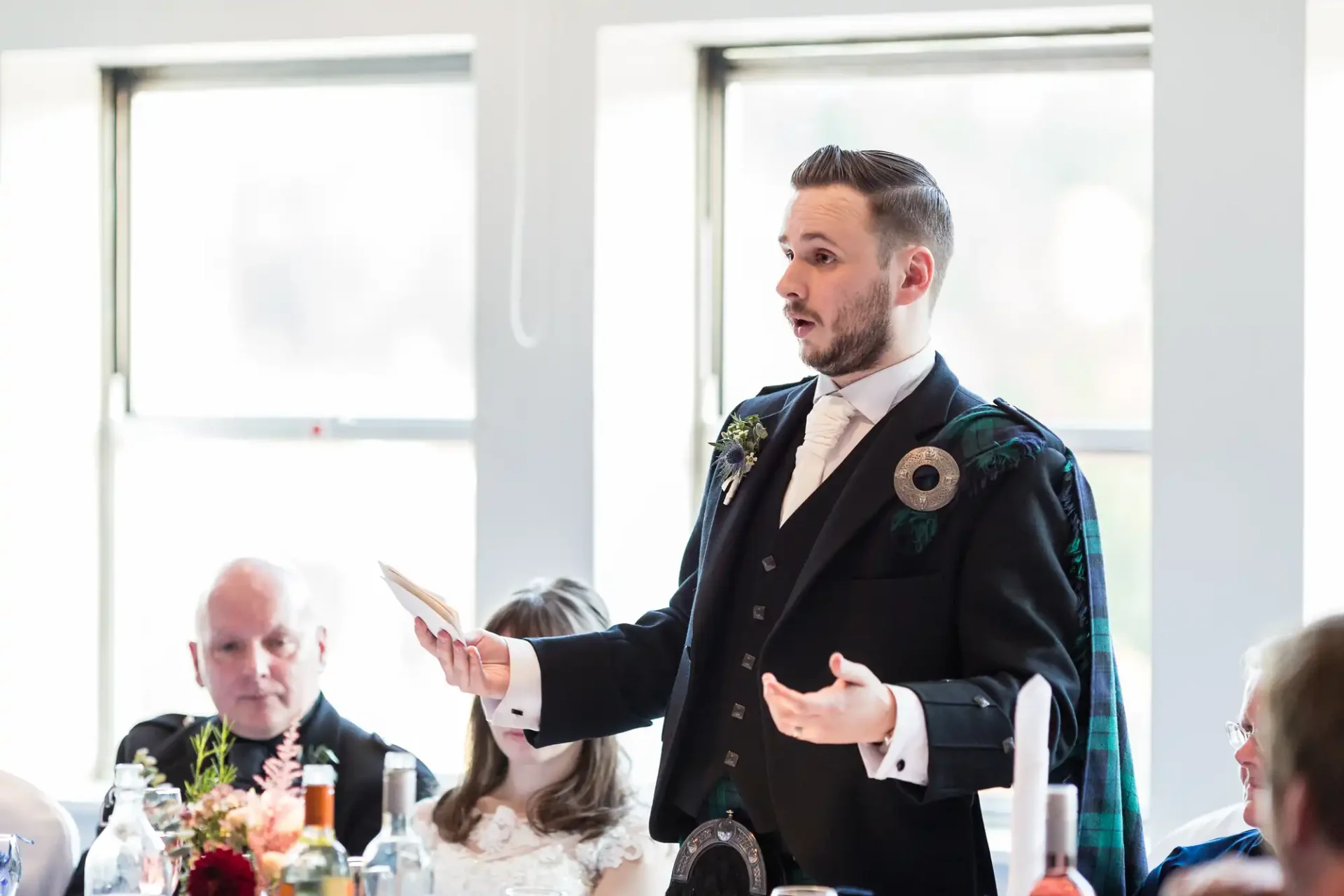 A man in a kilt and suit jacket giving a speech at a wedding reception, holding a note card, with guests seated around tables.
