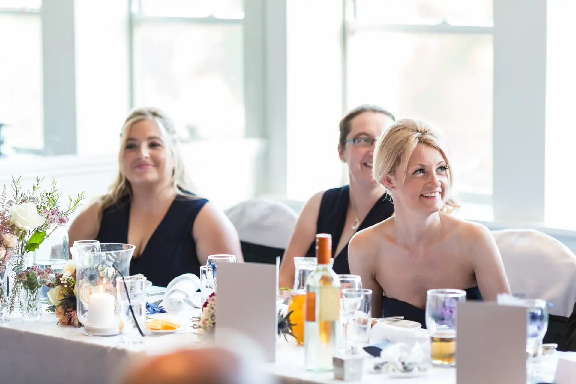 Guests at a wedding reception table, smiling and enjoying the event, with a focus on a blonde woman in the foreground. elegantly dressed with natural light streaming through windows.