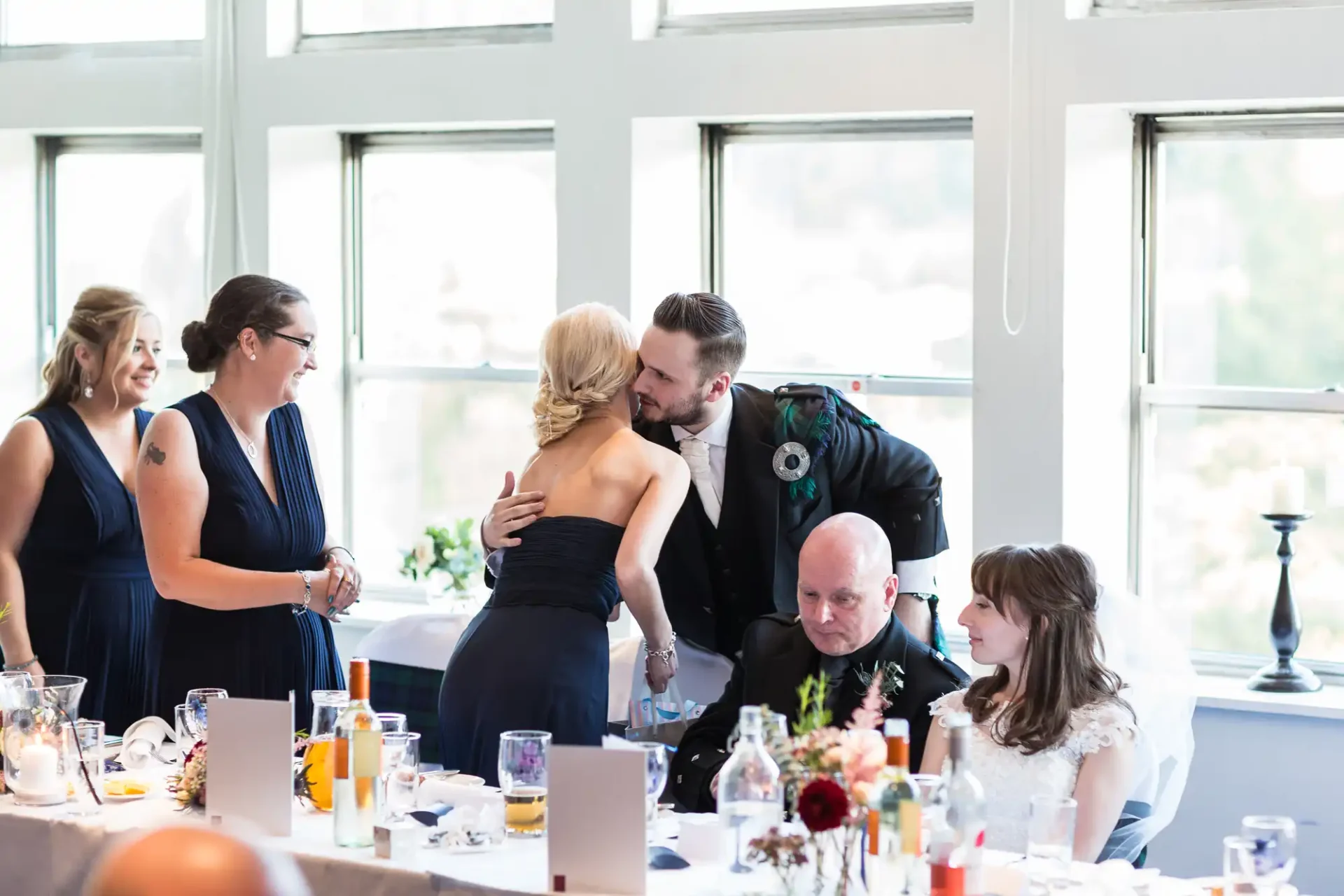 A man in a suit kisses a woman in a blue dress at a wedding reception, surrounded by seated guests and floral decorations.