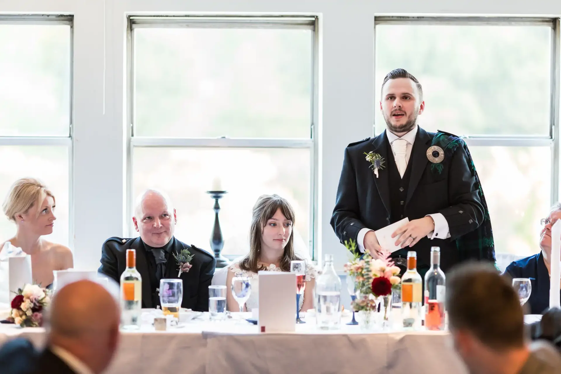 A man in a suit giving a speech at a wedding reception, surrounded by guests at a table.