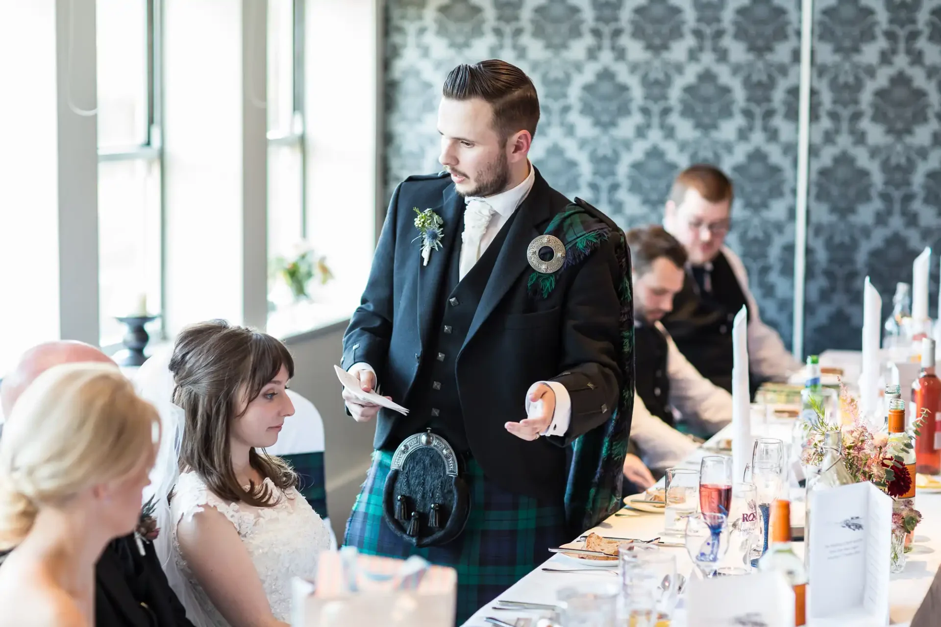 A groom in a kilt holding a microphone and speech notes at a wedding reception table, with a bride and other guests listening attentively.