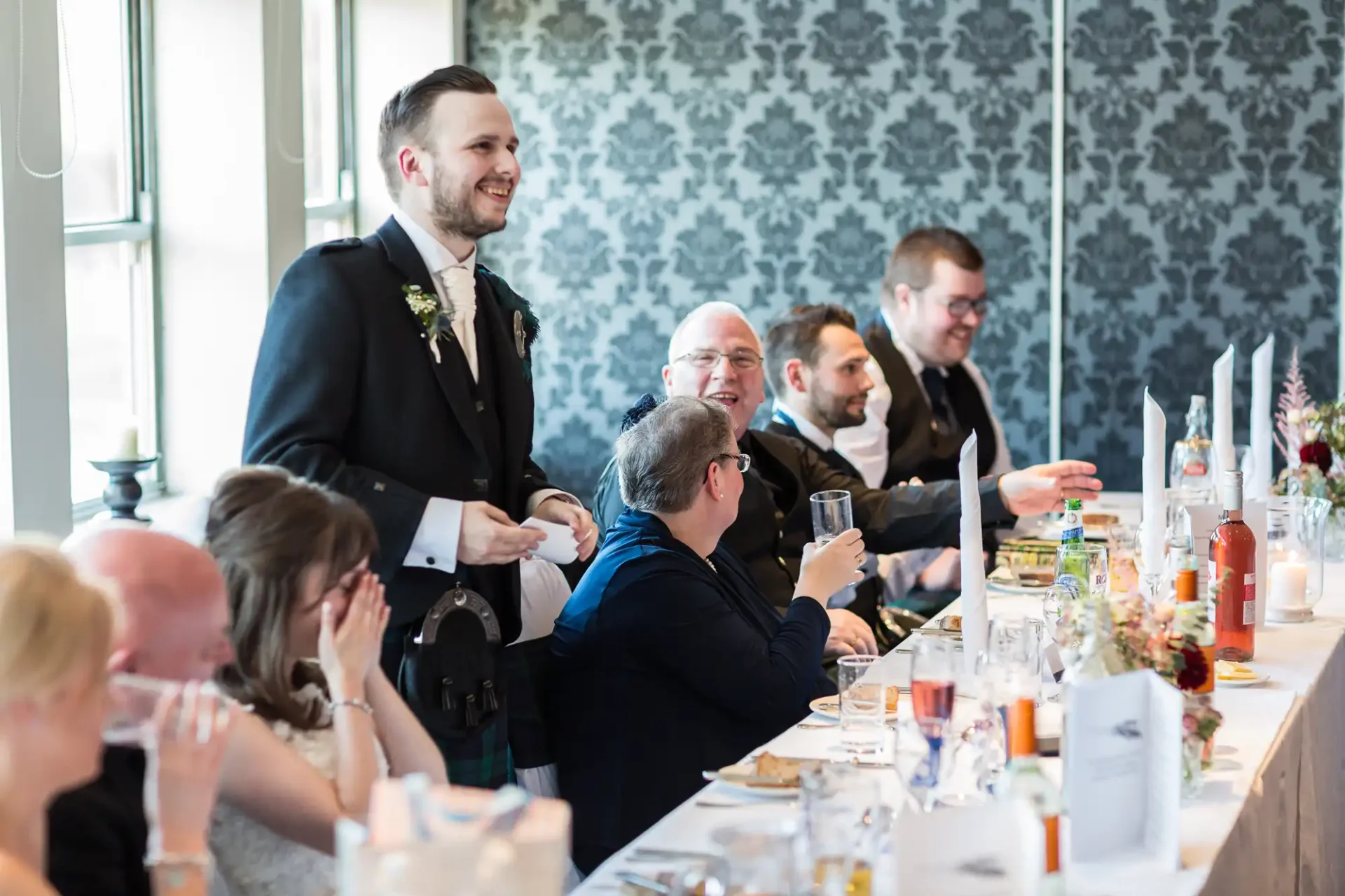 Groom smiling and standing beside seated guests enjoying a toast at a wedding reception table.