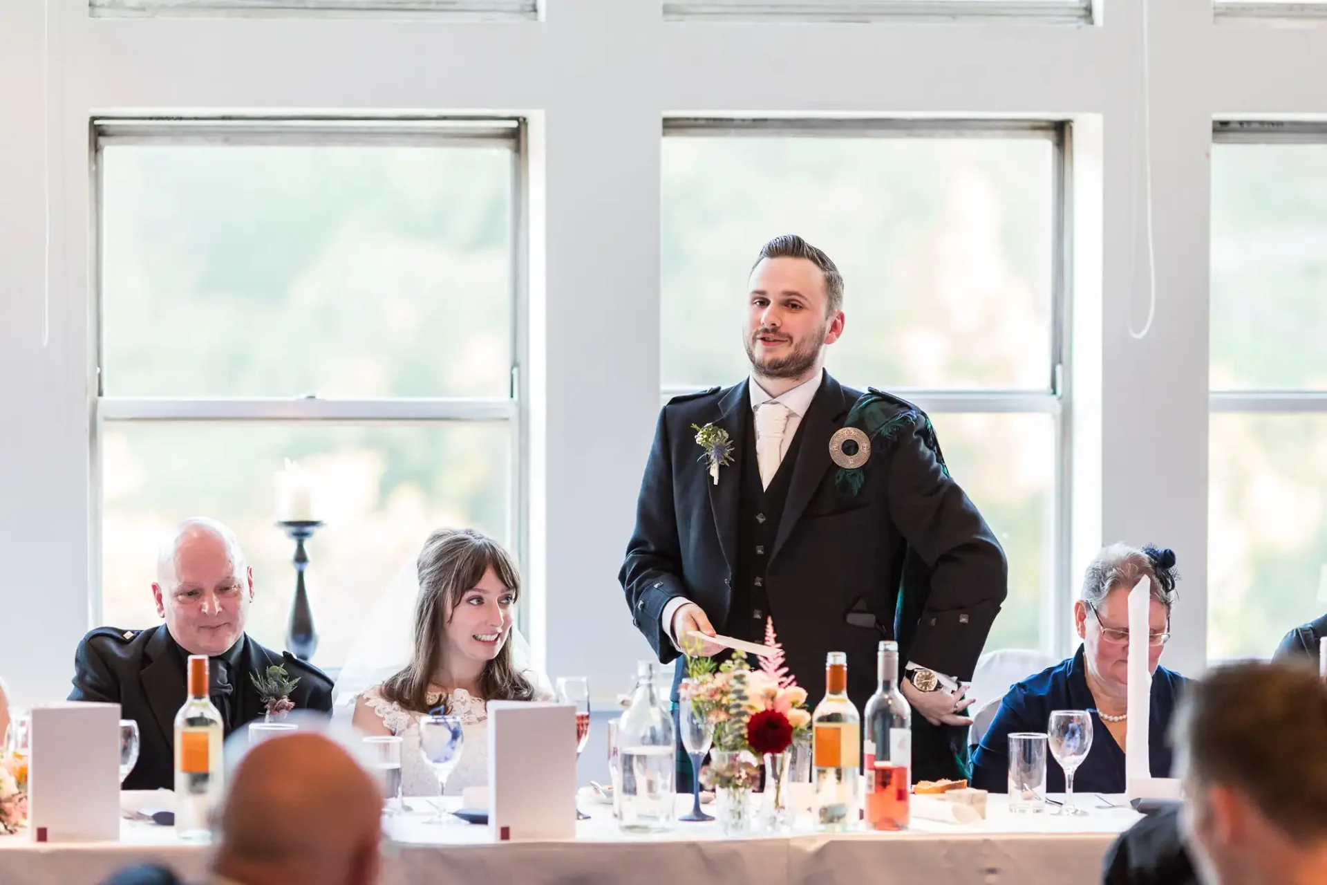 Man in suit standing and speaking at wedding reception table, with seated guests listening attentively.