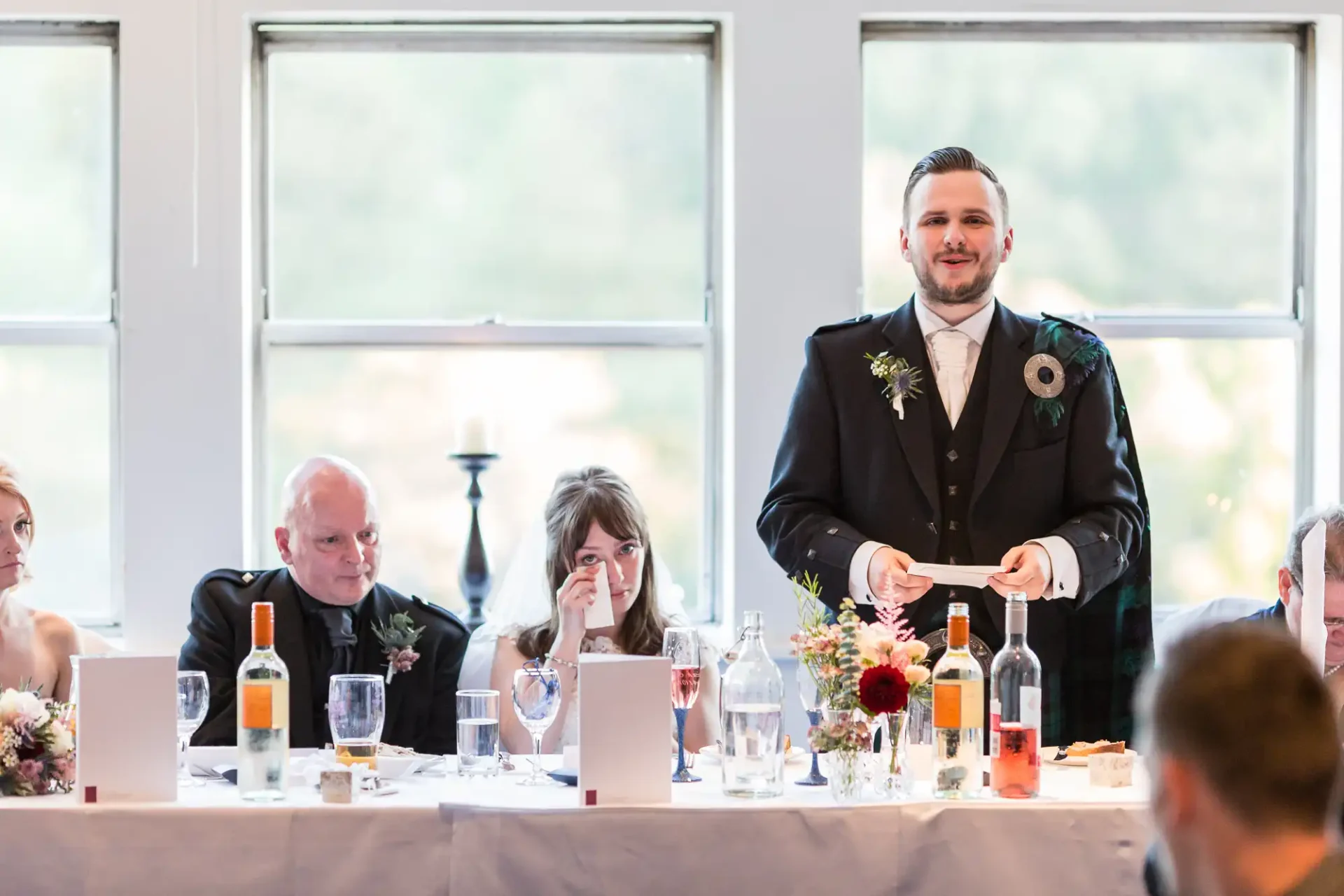 Groom standing and giving a speech at a wedding reception, with guests seated around tables listening.