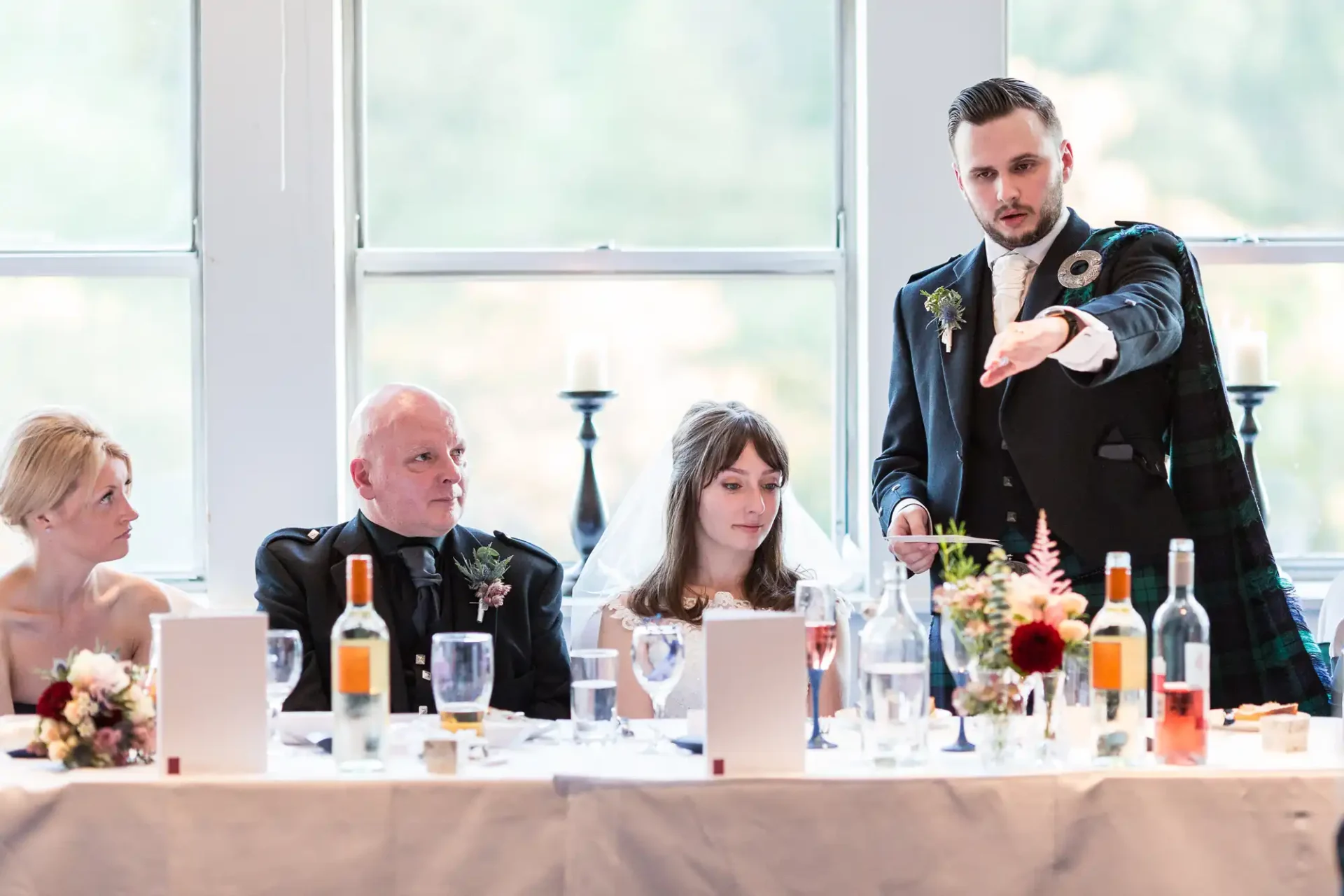 A man in a kilt speaks animatedly at a wedding table, pointing forward, while a bride and three other guests listen attentively.