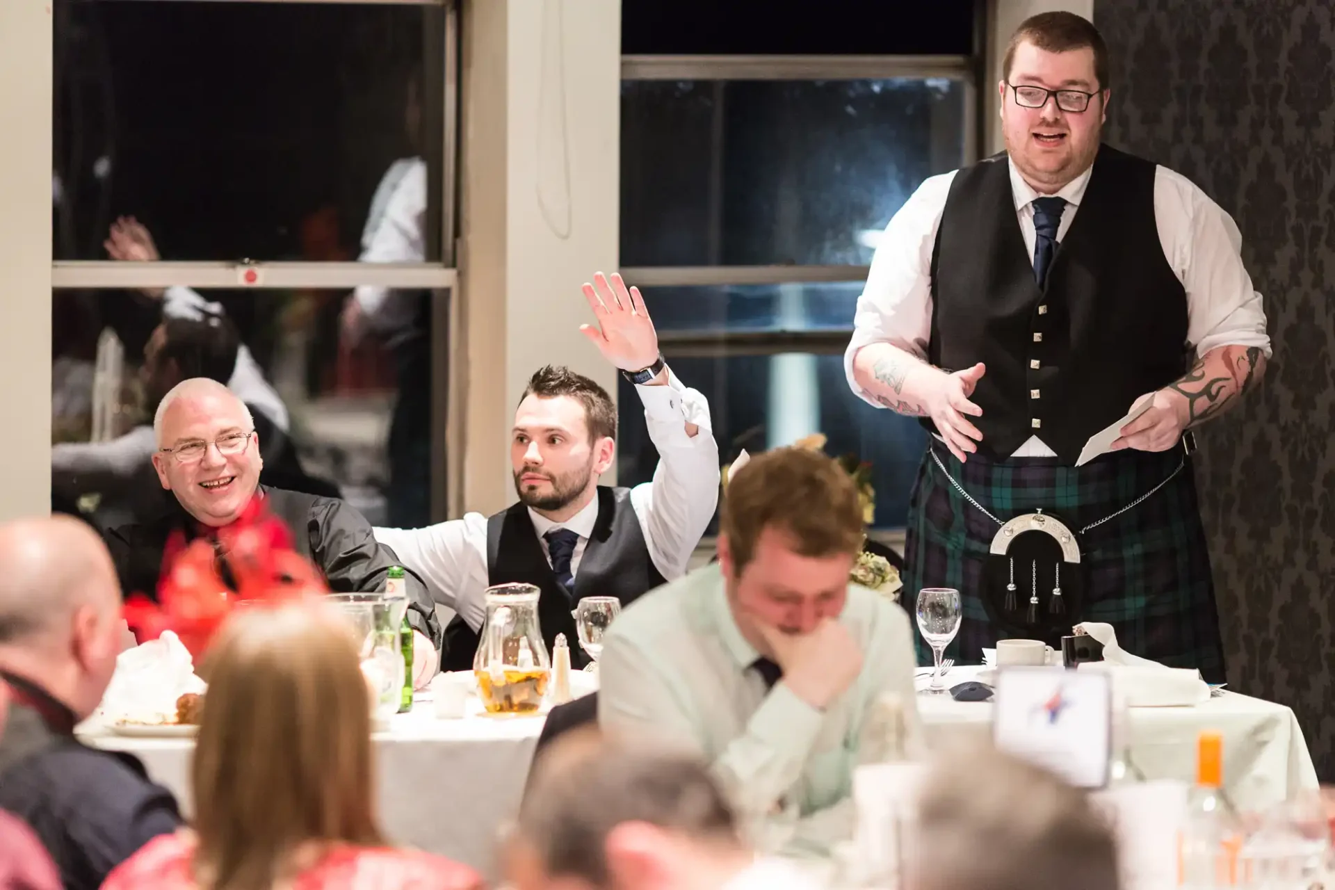 A man in a kilt speaks animatedly at a dinner event while guests around him listen and react, seated at tables adorned with festive decorations.