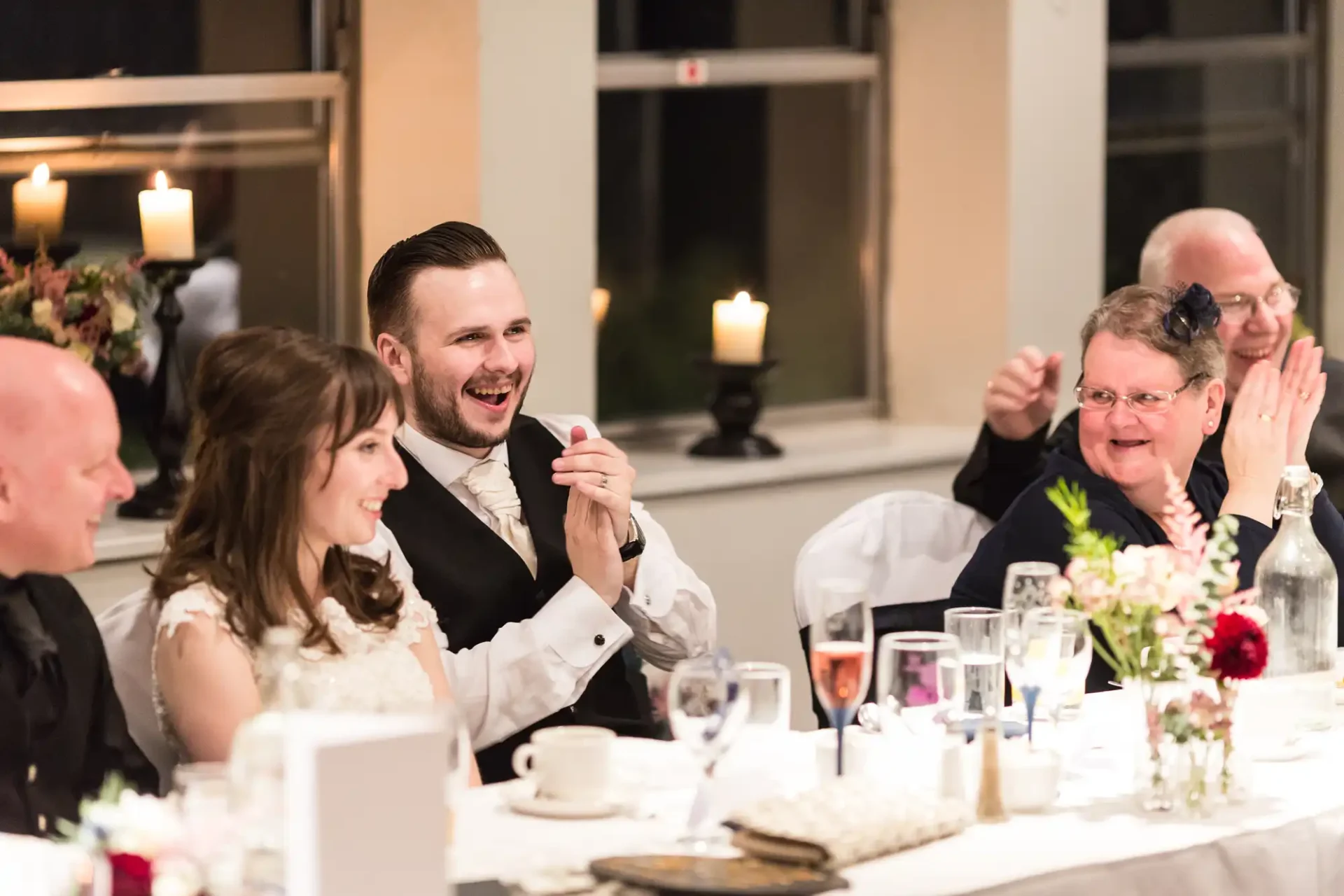 A bride and groom laughing at a wedding reception table, surrounded by joyful guests clapping and enjoying the evening.